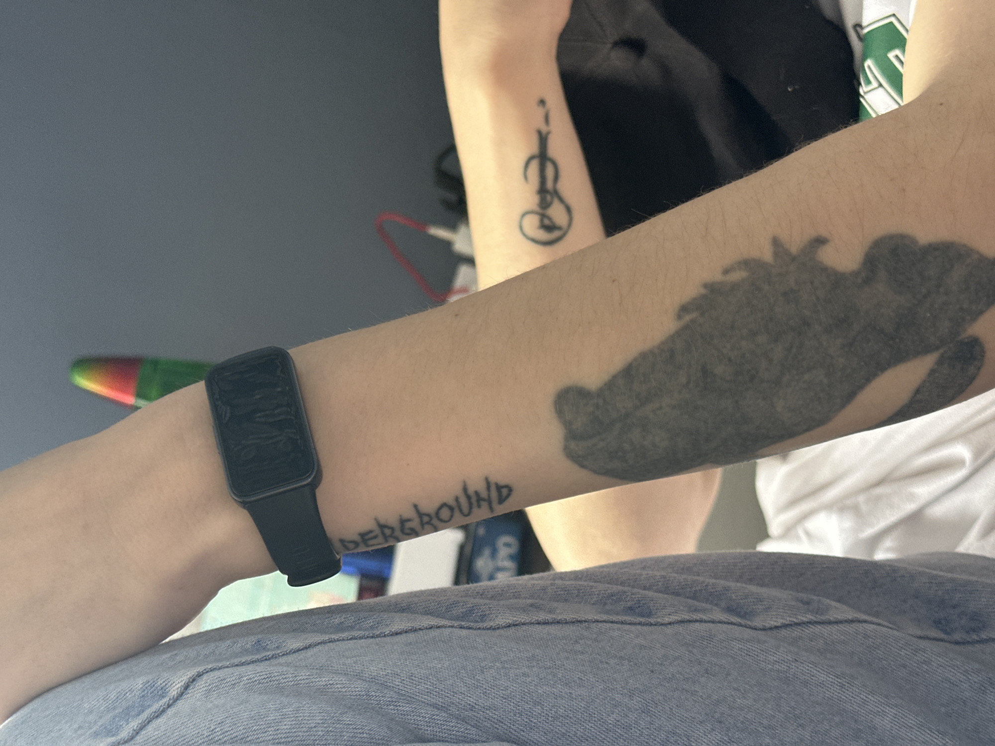 An image shows a partial view of two individuals, identified by their arms and clothing, against a gray wall backdrop. The focus is on the left arm of one individual, which has several tattoos: one of a figure resembling a violin on the forearm, a large shaded tattoo resembling a dinosaur on the upper arm, and the word "DERGROUND" tattooed near the wrist. A black smartwatch is worn on the wrist. The individual is wearing blue jeans. The second individual is wearing a black shirt and a white shirt with green lettering.
There is no indication in the image that it was taken in Stavropol, Russia.
