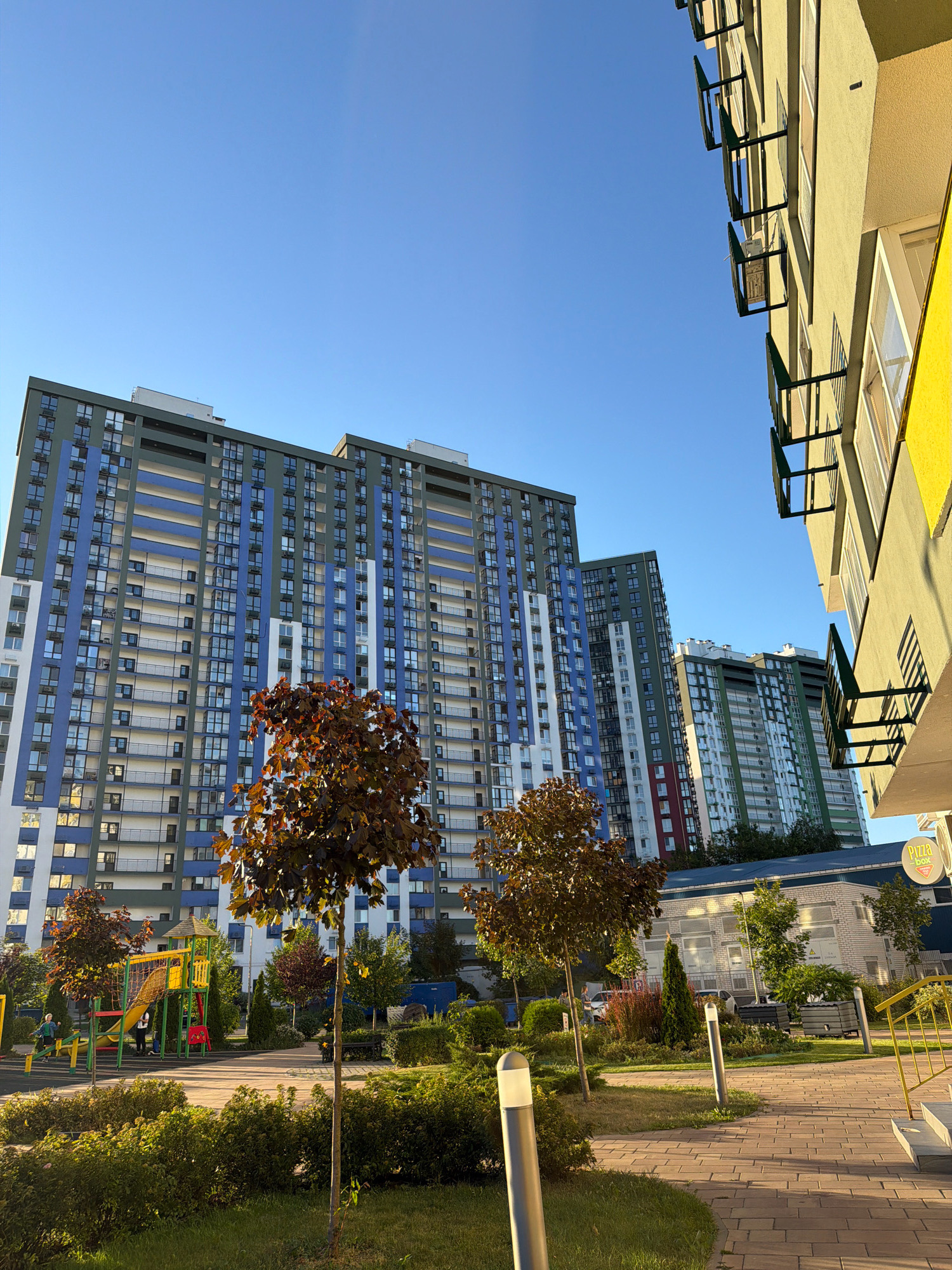 The image depicts a modern residential complex in Kyiv, Ukraine, under a clear blue sky. In the foreground, a paved walkway curves through a landscaped area with low green bushes and grass. Several young trees with reddish-brown foliage are scattered throughout the scene. To the left, a colorful playground features a slide, climbing structures, and a net, with two children visible.
Dominating the background are several tall apartment buildings with multi-colored facades, featuring balconies and numerous windows. The buildings are designed with a mix of blue, grey, white, and some red and green accents. To the right, a portion of a lighter-colored building is visible, also with windows and balconies. A yellow sign with the text "PIZZA BOX" is partially visible on this building. The overall impression is one of a new, well-maintained urban living space.
