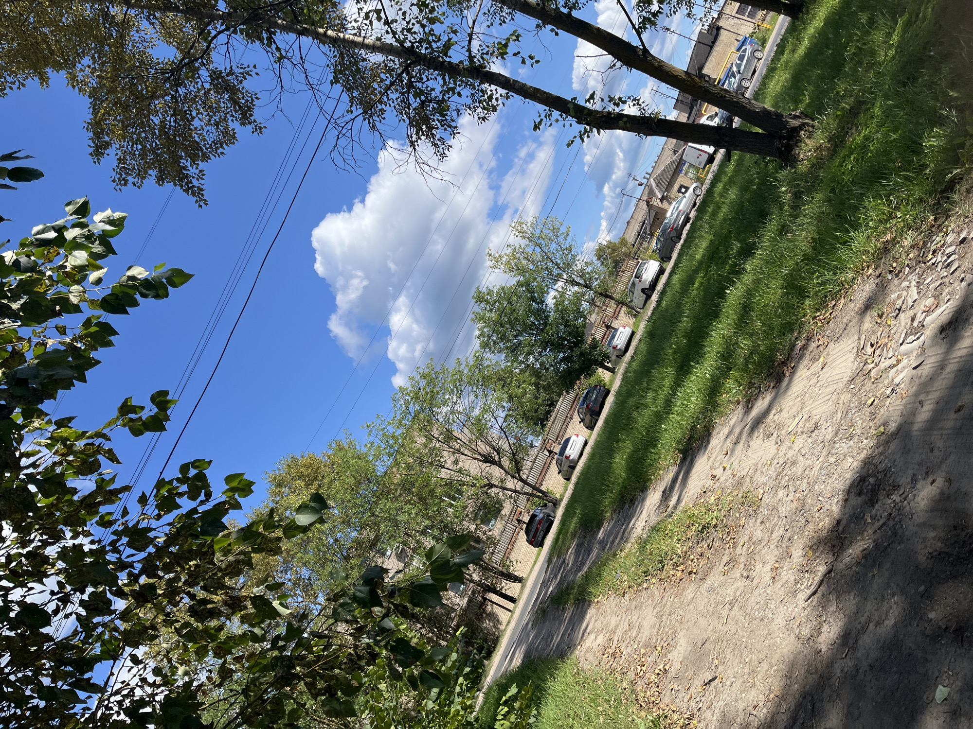 The image depicts a view from a dirt road angled upwards and to the right, bordered by a strip of green grass. Several cars are parked along the side of the road. In the background, a brick building with multiple windows is visible, adjacent to a chain-link fence. Trees with green leaves line the area, and power lines stretch across the bright blue sky, which is dotted with white clouds. The scene appears to be a residential or semi-industrial area within the city of Ivanovo, Russia.