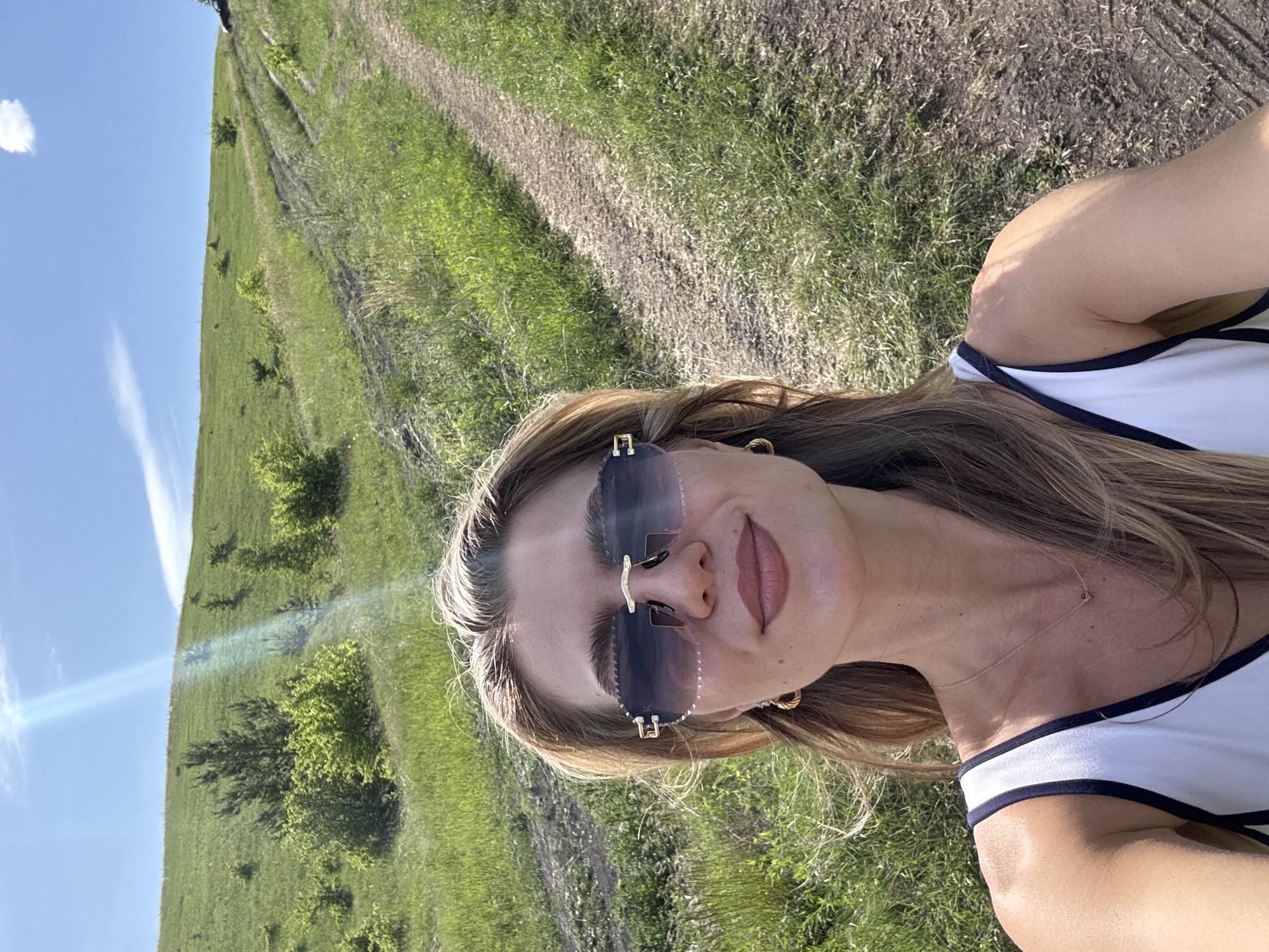 The image captures a selfie of a fair-skinned woman outdoors under clear blue skies with scattered white clouds. She has light brown hair, wears large, rimless sunglasses with gold accents and a scalloped upper edge, and a white tank top with dark trim. Her lips are closed in a slight smile, and she wears gold hoop earrings and a thin gold chain necklace with a small circular pendant. A prominent lens flare extends diagonally from the upper left. The background features a steep, grassy green hill dotted with small trees and bushes. A dirt path with tire tracks ascends the hill to the right, leading towards a dark-colored vehicle partially visible at the crest. The location is Unknown, Unknown.