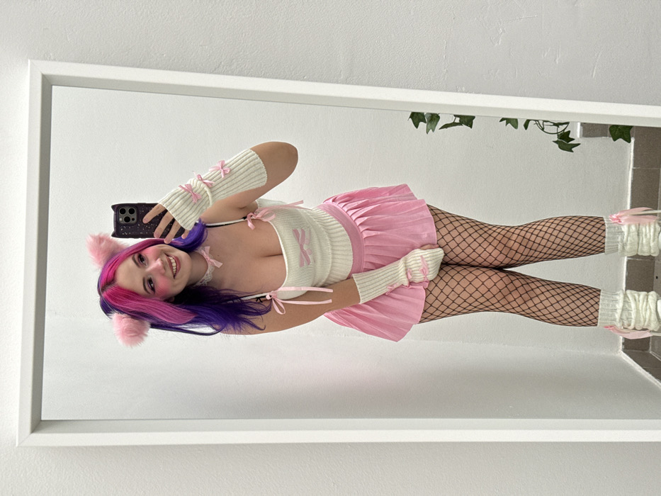 The image displays a young woman taking a mirror selfie indoors. The photograph is oriented sideways, with the woman and her reflection appearing rotated 90 degrees clockwise. She is smiling broadly and looking directly at the camera of her smartphone, which she holds in her left hand, visible in the reflection.
The woman has vibrant, multi-colored hair, transitioning from dark purple to bright pink, and is wearing fluffy pink cat ears. Her makeup includes noticeable blush. She is dressed in a "kawaii" or "e-girl" style outfit. Her top is a light-colored, possibly cream or off-white, ribbed or knitted camisole with thin straps. It features several small pink bows, with one larger bow centrally located on the chest and smaller ones on the straps. Around her neck, she wears a light-colored choker, possibly lace, also adorned with a small pink bow. Matching the top, she wears light-colored knitted fingerless arm warmers, each decorated with three small pink bows.
Her bottom half consists of a short, pleated pink skirt worn over black fishnet stockings. Completing her legwear are light-colored, ruffled leg warmers or cuffs around her ankles, which also have pink bows attached. The smartphone she is holding has a dark-colored case with a slightly textured or glittery appearance.
The setting appears to be a room with light-colored, possibly white, walls, reflected in a large mirror with a simple white frame. In the upper right portion of the mirror's reflection, some green foliage or artificial vines are visible, suggesting decorative elements in the room. The overall impression is one of playfulness and cheerfulness. No text is visible in the image.