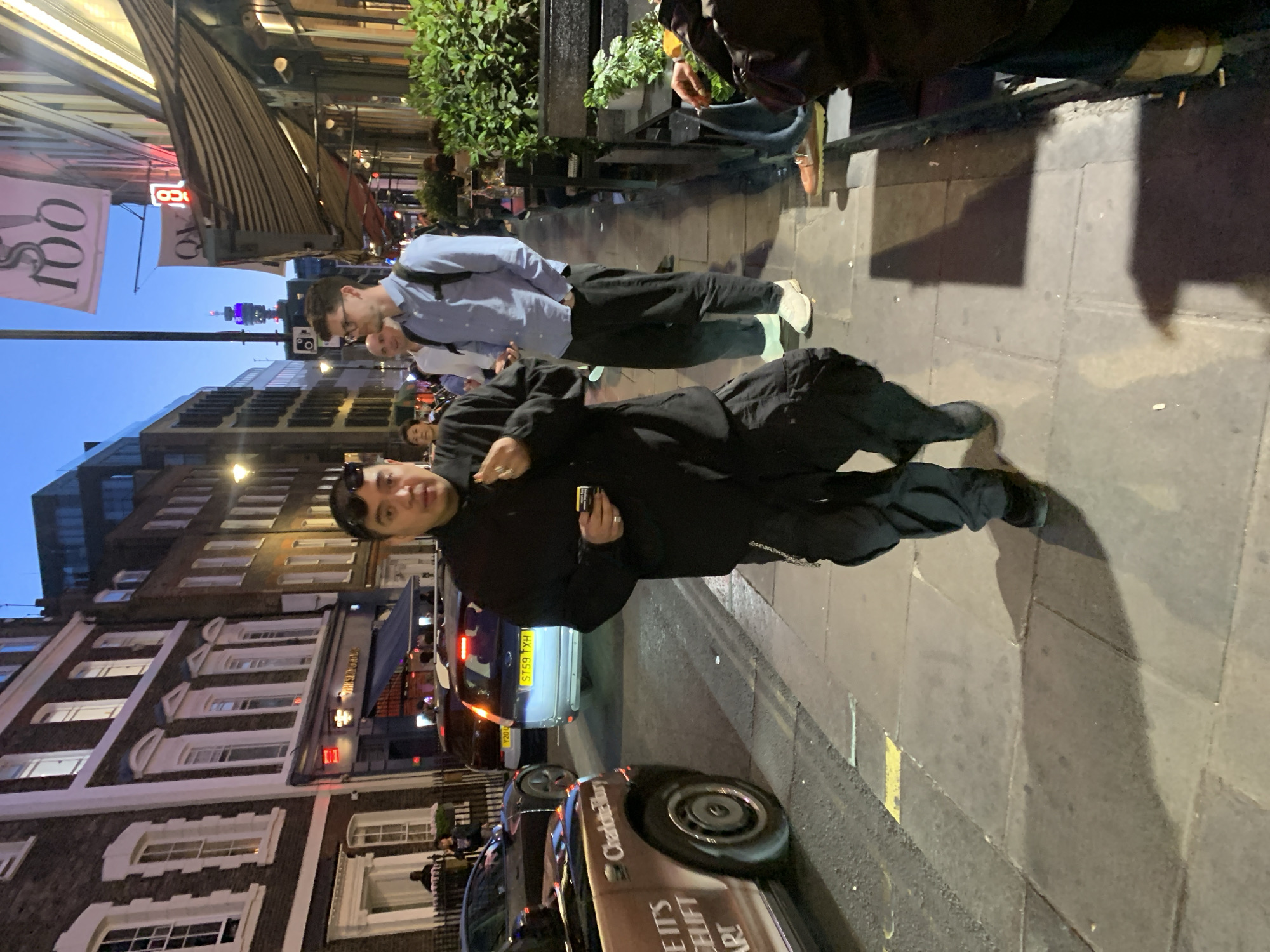 The image captures an evening street scene in an urban environment, likely London, United Kingdom. In the immediate foreground, a person with dark hair and sunglasses, wearing a black long-sleeved top and black trousers, looks directly at the camera while holding a small rectangular object. They are standing on a paved sidewalk.
Behind and to their right, another man in a light blue collared shirt, dark trousers, and white sneakers walks, looking downwards. Further back, more pedestrians are visible on the busy sidewalk, some near outdoor seating areas of businesses. The street is lined with multi-story brick and stone buildings, characteristic of London architecture, some featuring awnings over storefronts.
Artificial lights from streetlamps, building windows, and neon signs illuminate the scene, casting long shadows across the pavement, indicating dusk or early night. A light-colored car with a UK license plate "S789 TXH" is visible on the road in the mid-ground. In the lower foreground, part of a vehicle with "Charlottebury" and partially visible text "IT'S A LIFT ARE!" is seen.
Visible text includes a reversed "S 100" on a banner, a red neon sign displaying a reversed "QUO" and "B", and "The Sun Tavern" sign on one of the buildings. In the distant background, a tall structure with purple lighting stands out against the dimming sky, possibly a city landmark. The overall atmosphere suggests a lively city street as evening sets in.