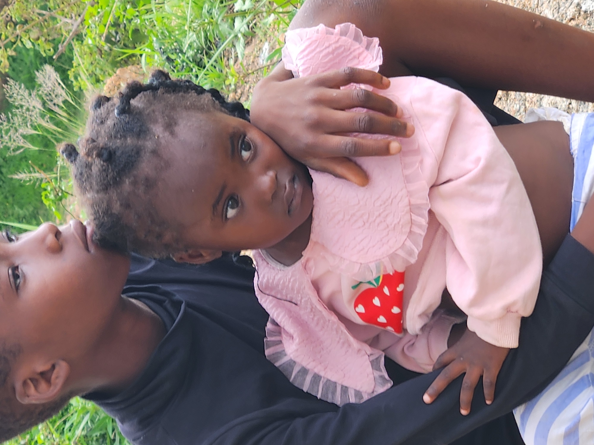 The image shows a close-up, portrait-style shot of two young Black children outdoors, likely in a natural or semi-natural setting. A slightly older child, appearing to be a boy, is holding a younger child, a girl, who is nestled against his chest. The older child is wearing a dark, high-necked long-sleeved shirt. The younger child is wearing a light pink ruffled top with a strawberry print on the front. The children's faces are visible, with the younger child looking slightly to the side. The background consists of blurred greenery, suggesting vegetation and possibly some sparse foliage, consistent with an outdoor environment in a city like Jos, Nigeria, which has a predominantly savanna climate with varying degrees of vegetation depending on the season and specific location. The lighting appears to be natural daylight.