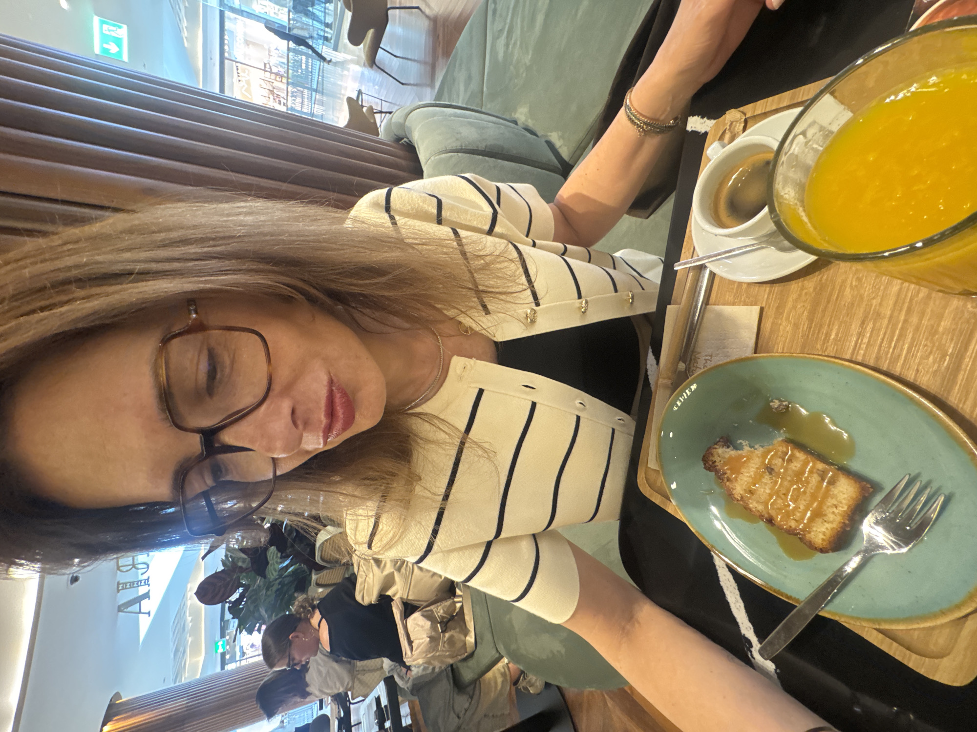 A close-up, selfie-style image captures a woman seated at an indoor café or restaurant in Senhora da Hora, Portugal. The image is rotated 90 degrees clockwise, showing the woman's face and upper body in the foreground. She wears brown-framed glasses and has light brown hair. Her attire includes a cream-colored cardigan with vertical black stripes, worn over a black top. A thin necklace is visible, and several bracelets adorn her right wrist.
On the wooden table in front of her are several items: a slice of cake, possibly a pound cake or similar, drizzled with caramel or a sweet sauce, presented on a teal plate with a fork. Next to it, there's a white cup containing dark coffee (likely an espresso) on a white saucer. A large glass jug filled with a bright yellow beverage, consistent with orange juice, is also present.
The background reveals a modern and comfortable interior. There's a soft green upholstered bench or couch, and walls feature horizontal wooden paneling. Other patrons can be seen seated in the mid-ground, and plants contribute to the decor. Bright indoor lighting illuminates the scene. Faint, rotated text that appears to be "ABBA" or "ARBA" is visible on a pillar in the background, along with a green exit sign. The overall scene suggests a relaxed daytime meal or snack.