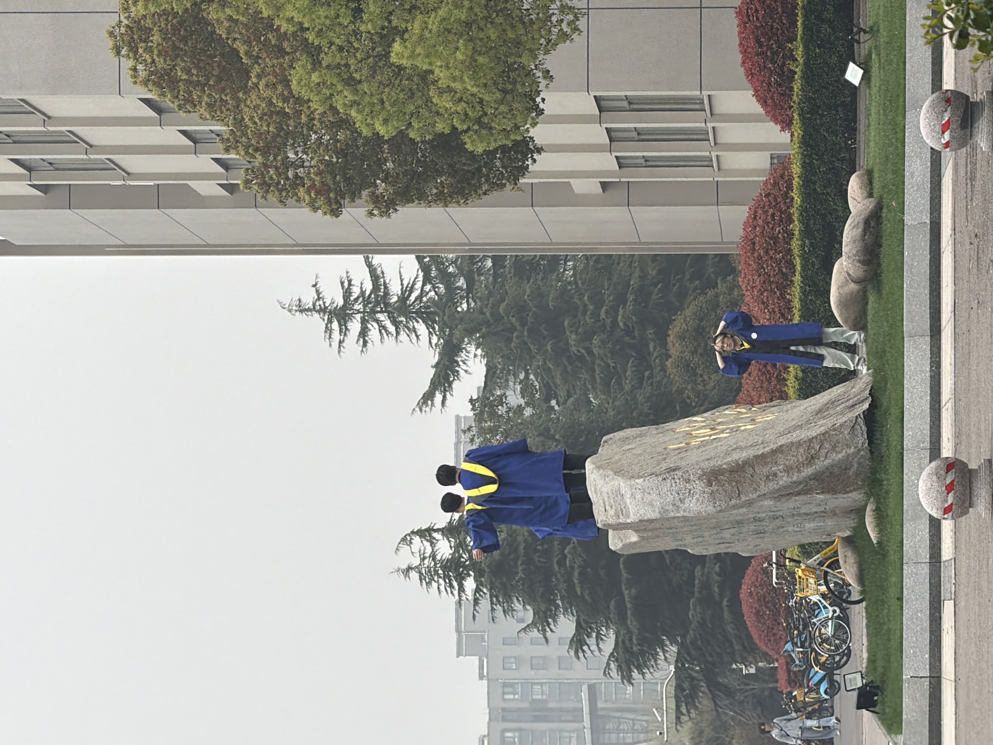 The scene is located in Jiangning District, China, near buildings and dense trees. Several individuals in graduation gowns stand near a large rock formation. A row of bicycles is parked close by. A person wearing a blue gown is standing on the grass near a row of bushes. There are stone markers on the right side of the image. The sky is overcast.