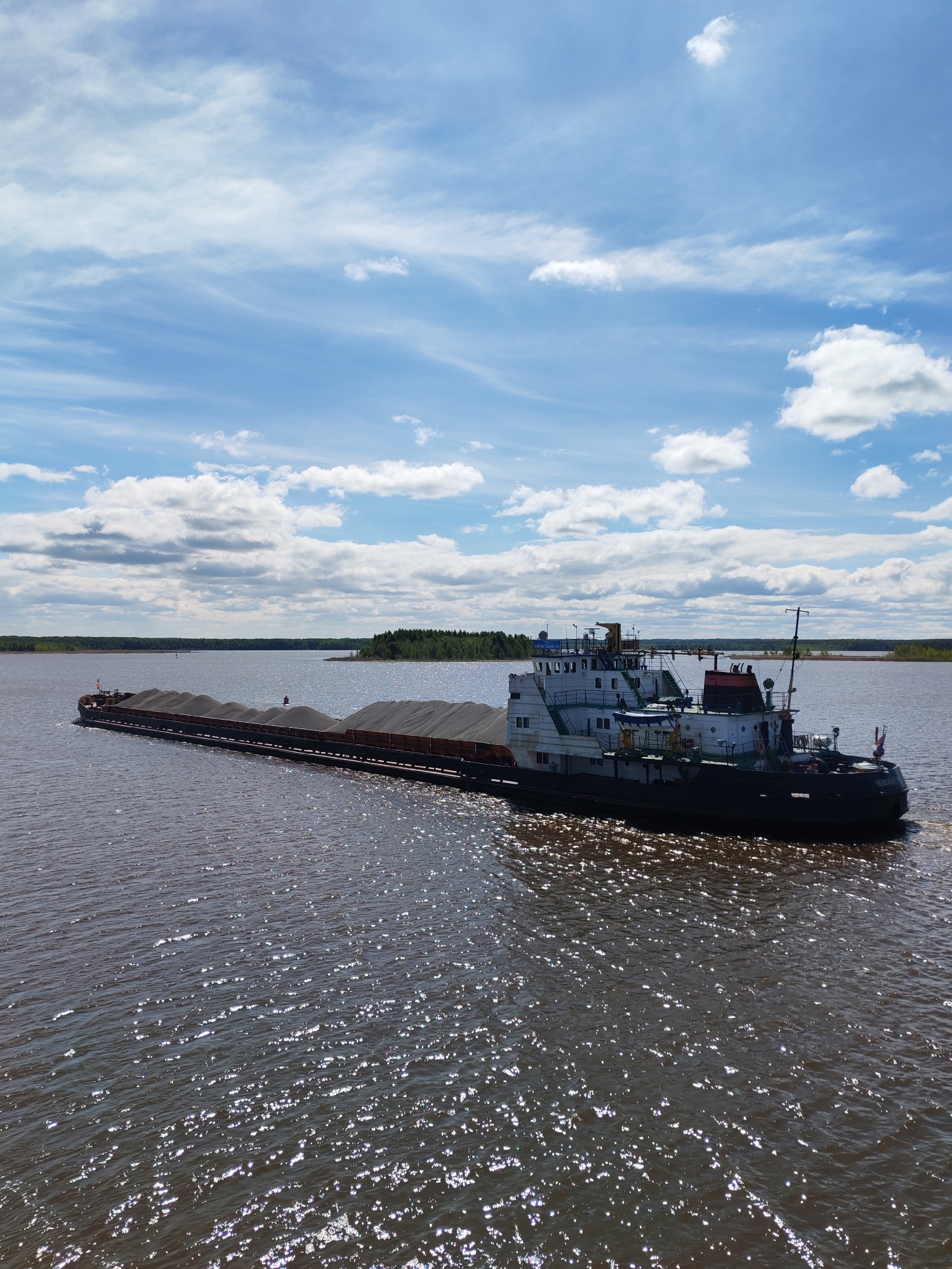A tugboat and barge traverse a body of water on a sunny day with a blue sky punctuated by cumulus clouds. The barge appears to be laden with a dark, granular material. In the distance, a shoreline with trees is visible. The water reflects sunlight, creating a shimmering effect. The city of Novodevich'e, Russia is not readily identifiable from this image.