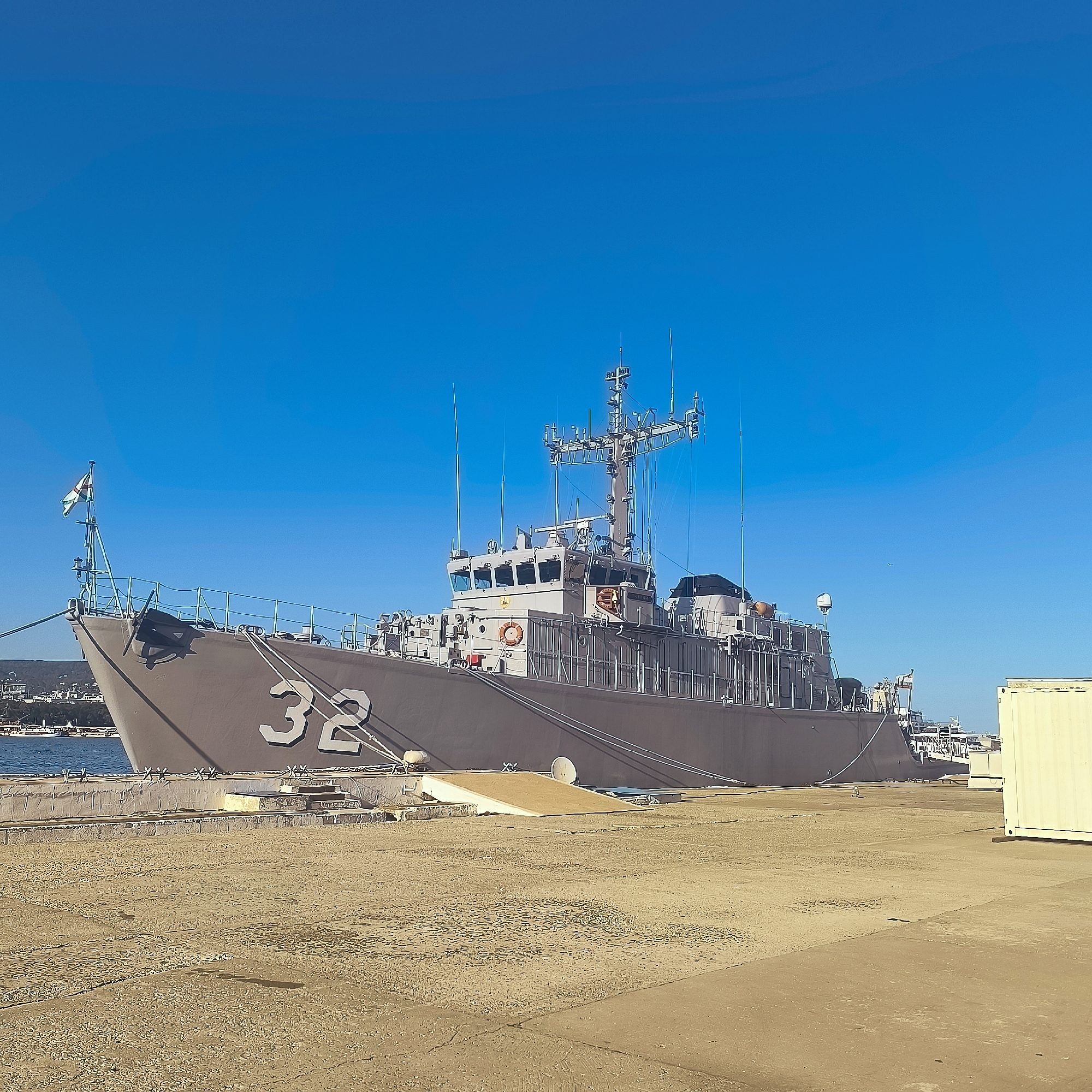 A naval ship, marked with the number "32", is docked at a pier in Varna, Bulgaria. The ship is gray with a light-colored superstructure, featuring various antennas and communication equipment. A flag with a white and green design is visible at the bow. The pier is made of concrete and dirt, with mooring lines connecting the ship to the dock. In the background, other ships and buildings can be seen along the coastline, under a clear blue sky. A yellow shipping container is also present on the pier to the right of the frame.