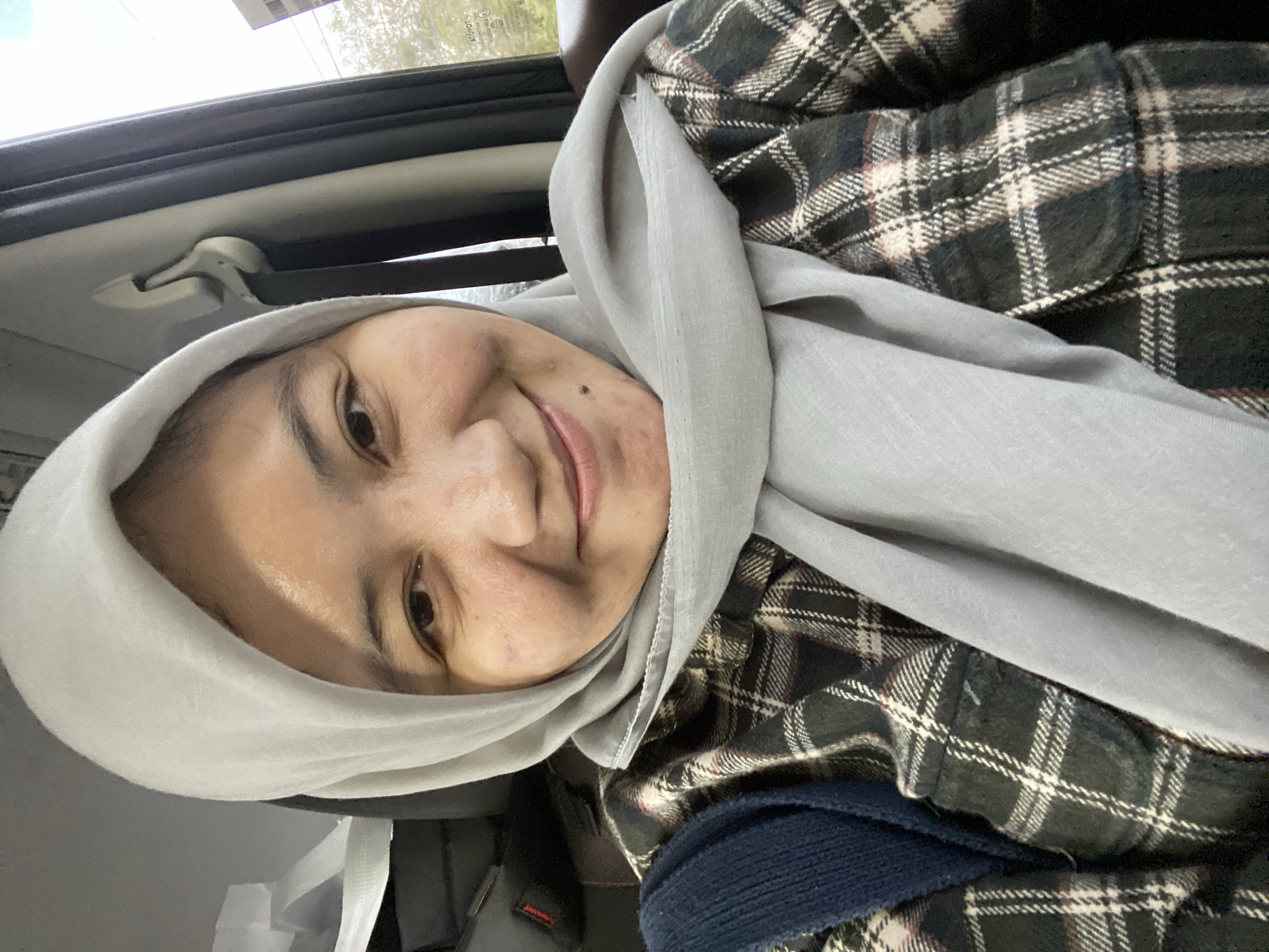 A close-up, selfie-style shot shows a young woman wearing a light grey hijab and a dark plaid shirt. She is in the passenger seat of a vehicle, with a grey seatbelt visible behind her. The background outside the car window shows trees and what appears to be a building. The scene suggests the woman is traveling within Banjarbaru, Indonesia.
