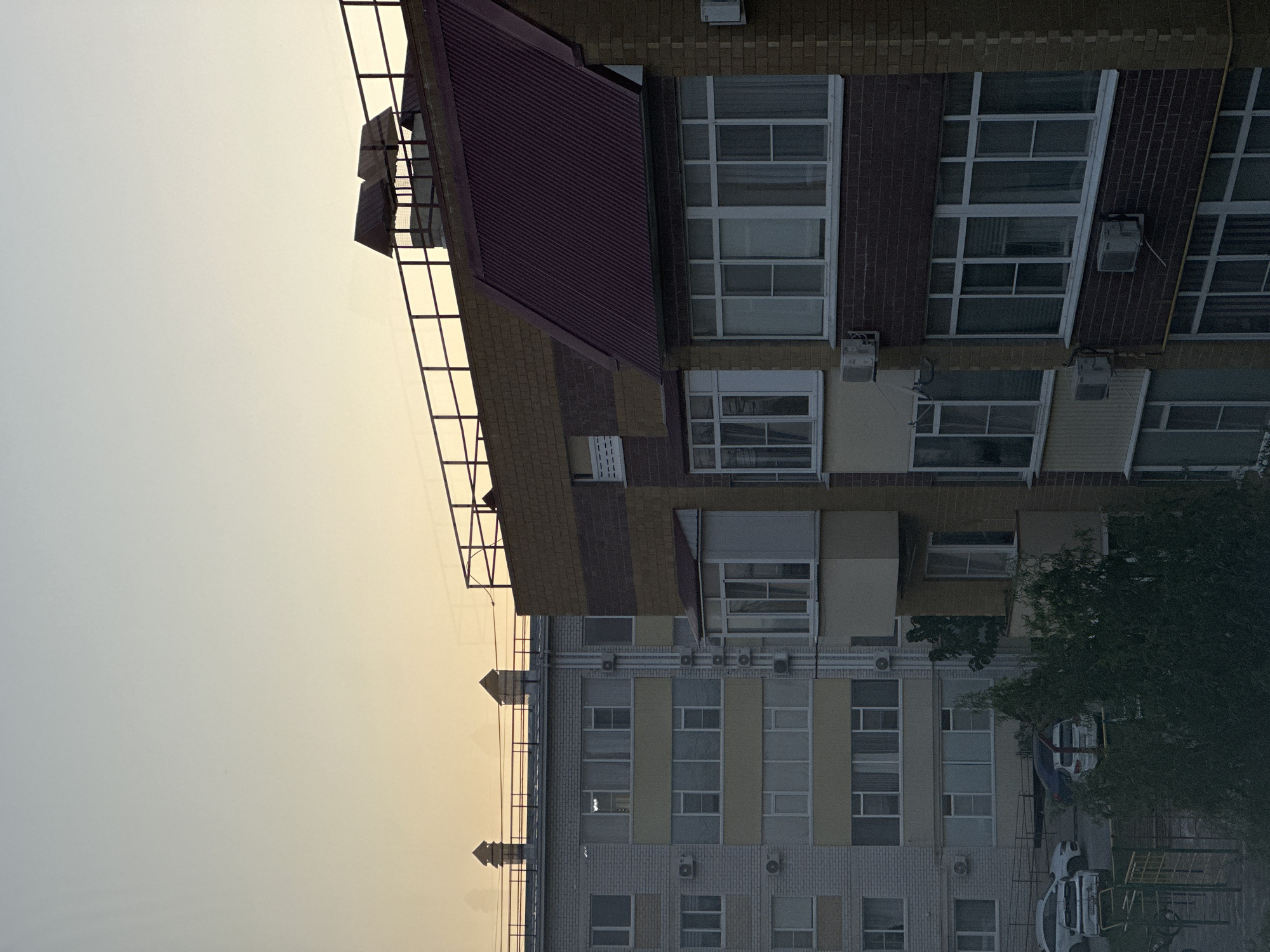 The image depicts a view of apartment buildings in Nevinnomyssk, Russia, under a pale, overcast sky that transitions from a light blue at the top to a warm yellow-orange hue near the horizon, suggesting either sunrise or sunset. The architecture is consistent with post-Soviet era multi-story residential buildings, characterized by light-colored brickwork and distinct balcony structures.
On the left, a lighter-colored building with multiple floors is visible, featuring numerous windows and balconies, some with yellow accents. The facade is a mix of white and pale yellow brick. Several air conditioning units are mounted externally on this building.
To the right, a taller building with a distinctive red-pitched roof dominates the central and right portions of the frame. Its facade is a combination of dark red and yellow brick. This building also has multiple windows and balconies, with some enclosed with glass. A dark metal railing runs along the top of the buildings.
In the foreground, the tops of trees are visible, with lush green foliage partially obscuring the lower levels of the buildings and the ground. A playground structure, possibly with yellow and green components, is also partially visible at the bottom left. Several cars are parked in the area between the buildings and the playground, including a white SUV and a dark-colored sedan. The overall impression is a typical urban residential scene.