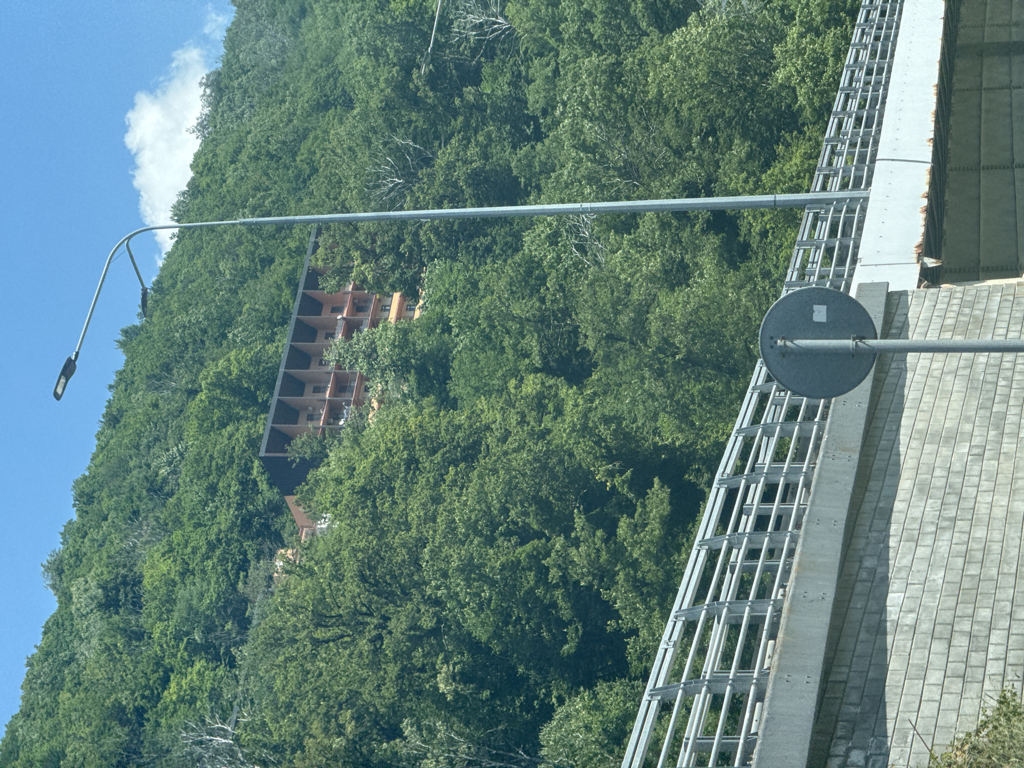 A multi-story building with a reddish-brown facade and dark, recessed balconies or architectural features is situated on a heavily wooded hillside. The building is partially obscured by the dense green foliage of numerous trees. In the foreground, a modern bridge structure with a grey concrete support wall and a metal railing is visible. A grey metal pole supporting a round, grey sign with a white sticker is positioned in front of the bridge. A tall, slender lamppost with two light fixtures arches over the scene. The sky above is a bright blue with scattered white clouds. The overall scene suggests a location in or near Krasnaya Polyana, Russia, characterized by mountainous terrain and significant vegetation.