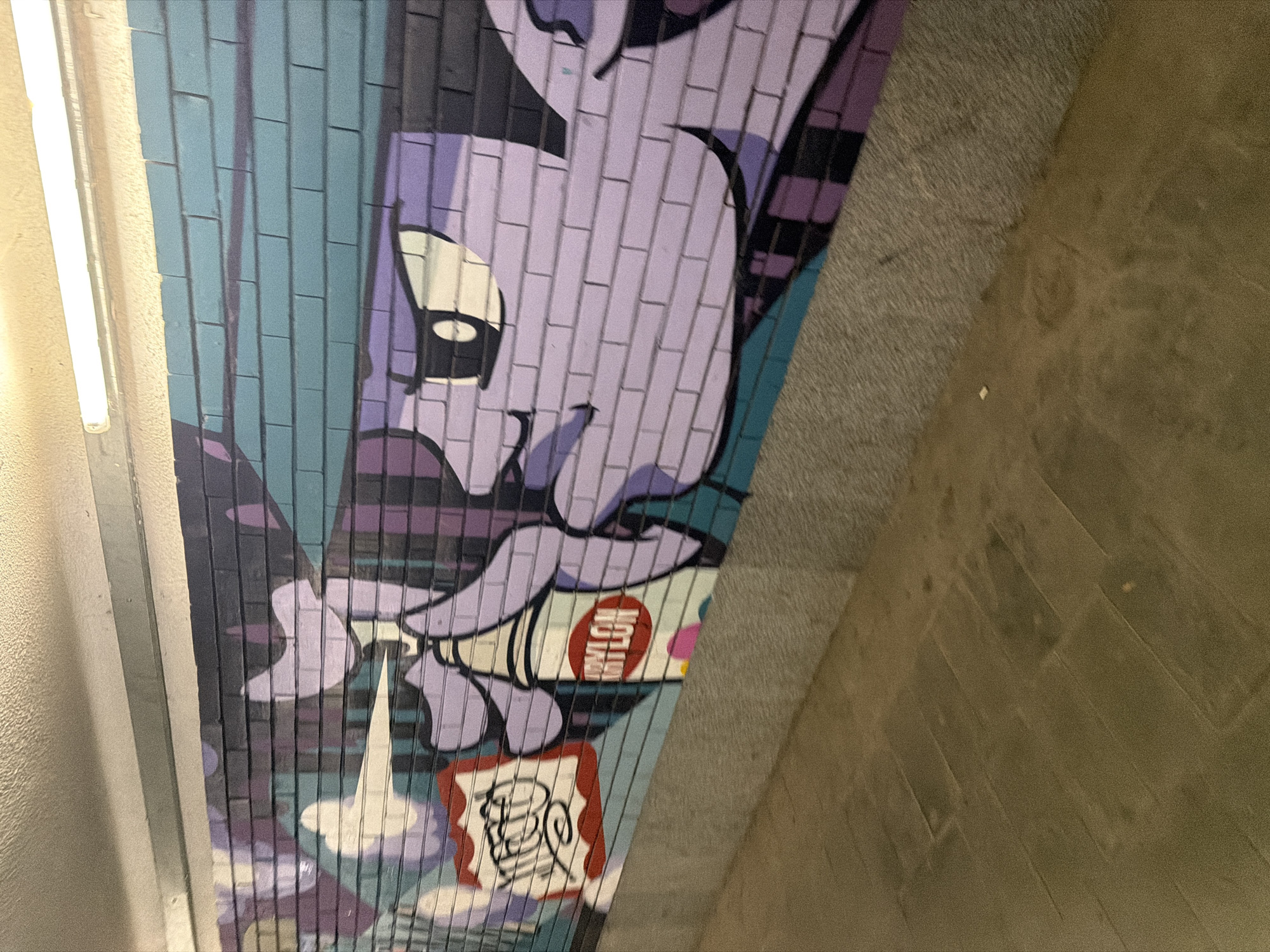 A graffiti mural on a brick wall depicts a cartoon character with purple skin and large eyes, wearing a teal hat. The character is holding a spray can with the brand name "MYLON" visible on it. The mural is illuminated by fluorescent lights above, and the lower portion of the image shows a concrete floor.