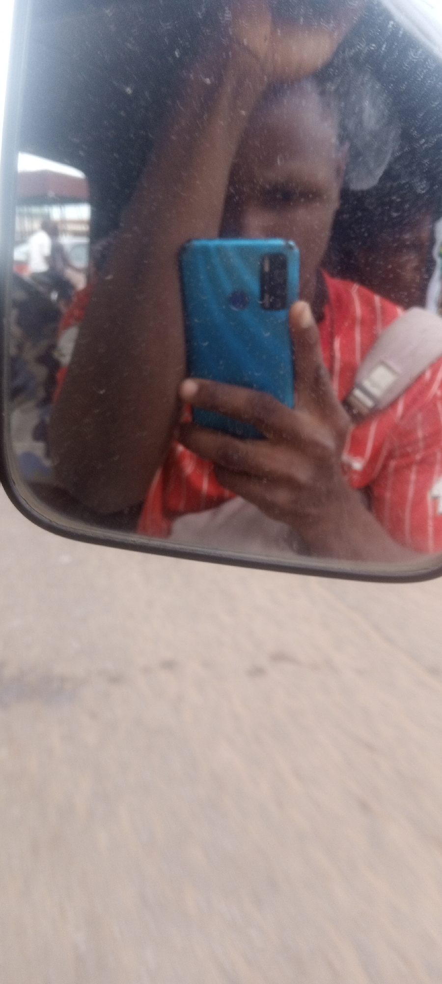 A selfie reflected in a side mirror shows a person holding a blue smartphone, presumably taking the photograph. The person is wearing a red striped shirt and has their hand on their head. The mirror reflects a partial view of the interior of a vehicle and the background appears to be a street scene, suggesting the location is in Sabon Tasha, Nigeria. There is a road below the mirror and people and vehicles can be seen in the blurred background, indicative of a bustling urban environment.