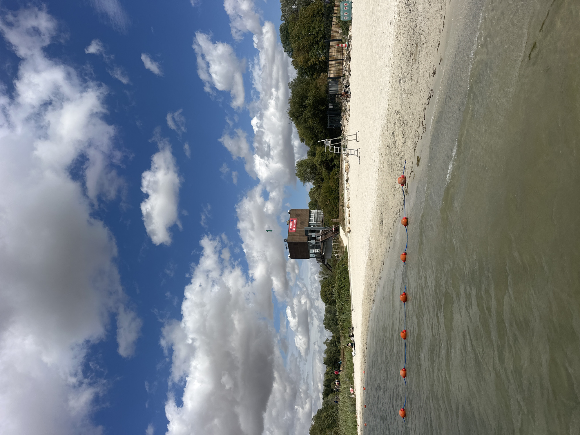 The image depicts a sunny day at the beach in Jablines, France. The foreground shows the edge of a body of water with a line of orange buoys marking a boundary. The water is calm and appears slightly murky. Behind the buoys is a sandy beach, with a few scattered pieces of seaweed.
Further back on the beach, several people are visible. On the left side, a small group of individuals are standing and walking, some appearing to be engaged in activities near the tree line. In the center-right, a lifeguard chair stands on the sand, with a person seated at its base.
Dominating the middle ground and background is a multi-story building that appears to be a lifeguard station or a recreational facility. It has a brown facade and a red sign on the upper level that reads "POSTE DE SECOURS" (Rescue Station). A green flag flies from the roof. Behind the building and extending to the sides is a dense line of green trees and foliage. The sky above is a vibrant blue, dotted with fluffy white and grey clouds. The overall impression is one of a recreational area on a pleasant day.
