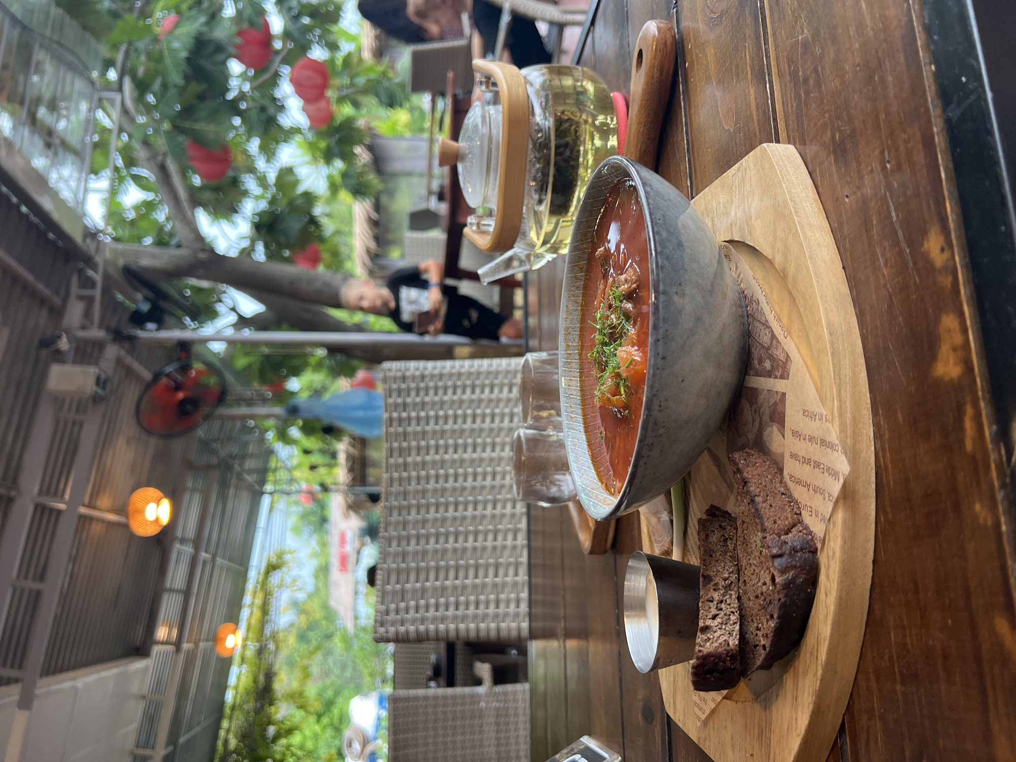 Here is a detailed description of the image:
**Scene Overview:**
The image shows a close-up view of a table setting, likely in an outdoor or open-air restaurant in an unspecified city. The orientation of the shot is turned sideways.
**Table Setting:**
The table is wooden with a visible grain. A round, wooden platter or board holds a bowl of red soup with garnish, possibly borsch, accompanied by dark, sliced bread and a small metal container of what appears to be cream or sauce. There is a transparent glass teapot and glasses next to the soup. The platter appears to have a newspaper article as a base underneath the food.
**People and Interactions:**
There are blurred figures of people in the background, including someone seemingly resting on a chair or lounger. The presence of the food and drink suggests that someone is about to or has just eaten a meal at the restaurant.
**Environment:**
The setting seems to be in a restaurant with both indoor and outdoor seating. A red fan is visible.There is greenery in the background, indicating an outdoor area or patio, with hanging decorations that resemble red lanterns or flowers. The lighting suggests it could be daytime or early evening, with light coming through the foliage.