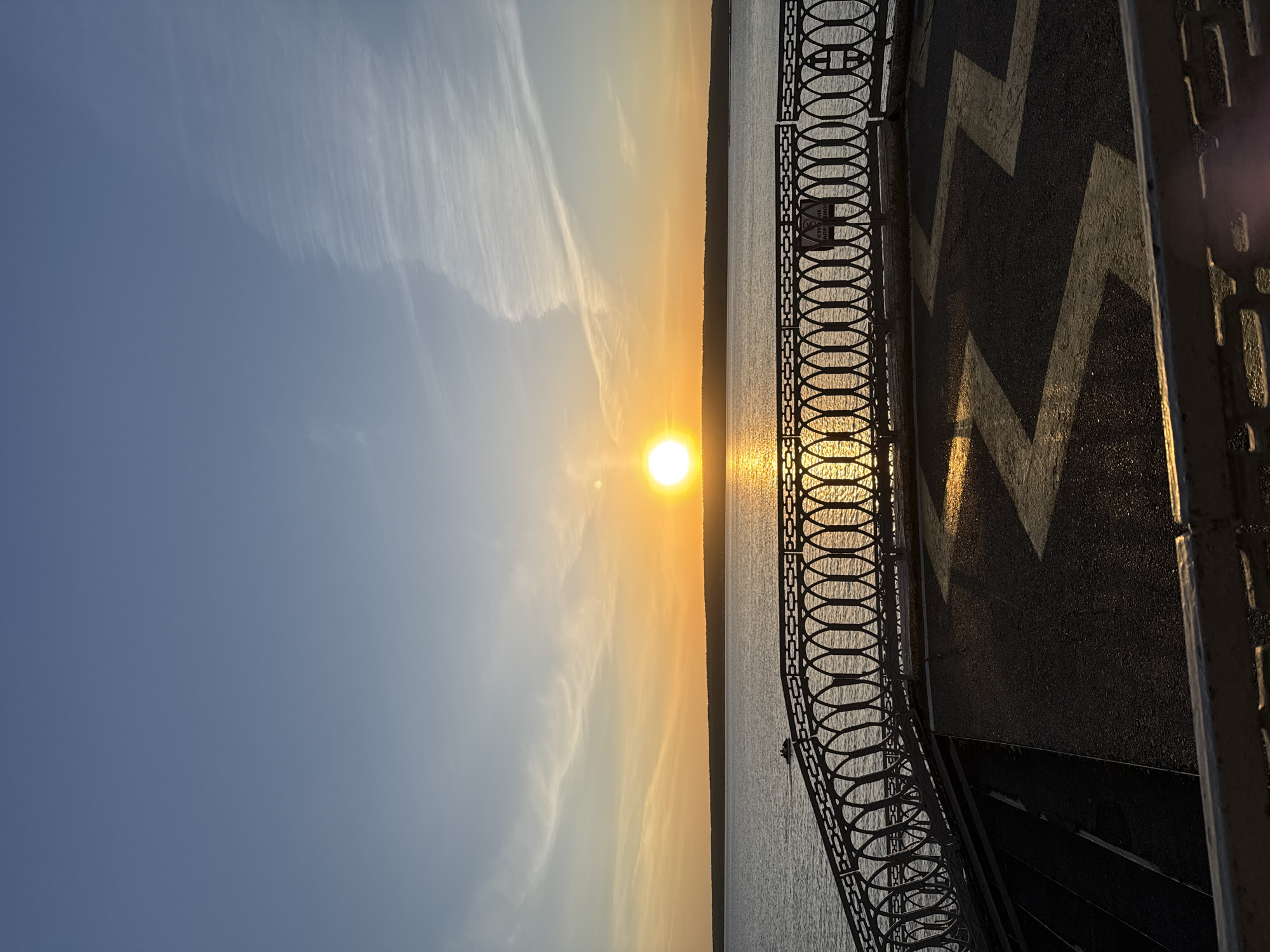 The image depicts a sunset over water, likely a river or large lake. The sun is positioned in the center of the frame, casting a reflection on the water surface. The sky is a gradient of blue to orange, with wispy clouds visible. A metal railing runs along the right side of the image, with a walkway behind it featuring painted chevrons. The location is Unknown, Unknown. The scene is devoid of people.