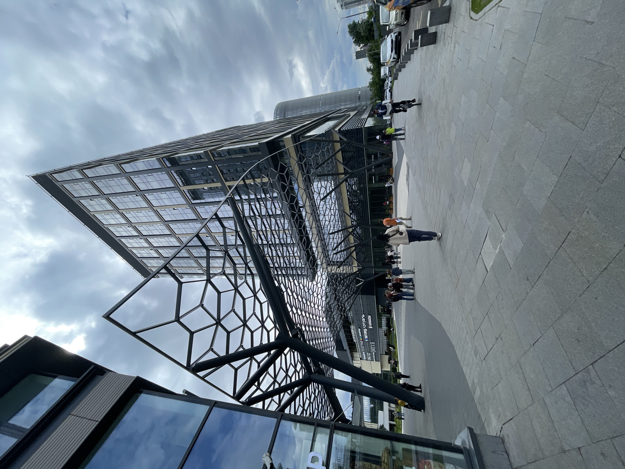 This media file captures a wide shot of a modern urban scene, predominantly featuring the exterior of a striking architectural building. The building exhibits a unique design with a large, angular glass roof supported by a complex geometric metal framework. This framework, composed of interconnected hexagonal shapes, creates an eye-catching visual element. The building itself appears to be made of glass and dark metal, reflecting the cloudy sky and surrounding environment.
In the foreground, parts of another building with large glass windows are visible, suggesting a commercial or public space. A paved plaza with large grey stone tiles stretches out in front of the main building, leading towards a street with parked vehicles, including cars and a white van. Several people are scattered across the plaza and sidewalk. Some are walking, while others are gathered in small groups. One person is riding a bicycle. The overall impression is a moderately busy day in a developed urban area.
The sky is overcast, with dense grey clouds dominating the upper portion of the frame, suggesting a potentially cool or damp day. The lighting is diffused, typical of an overcast sky, and there are no strong shadows. The time of day is indeterminate due to the lack of direct sunlight.
Visible text includes signage on the building's facade, although much of it is blurred or partially obscured. One sign appears to have English text, with words like "STUDIO COFFEE" and "ENTER" discernible. The architectural style and the presence of such businesses suggest a contemporary metropolitan setting. The people's attire is casual, further reinforcing the impression of an everyday urban environment. No specific emotions are readily apparent from the distant figures.