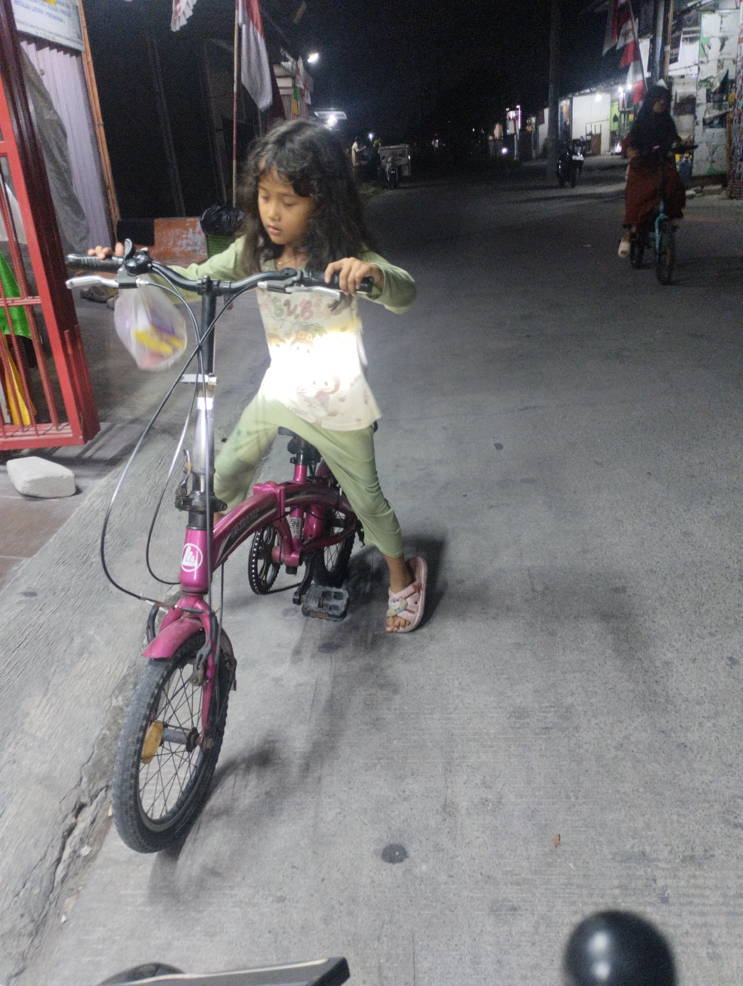 A young girl with long, dark, curly hair rides a pink foldable bicycle at night on a paved street in Karangraharja, Indonesia. She wears a light-colored shirt with a graphic print and green leggings, along with pink sandals. Her hands grip the handlebars, and she appears to be looking forward. The bicycle has a plastic bag hanging from the handlebars. In the background, another person on a bicycle is visible in the distance, along with buildings and streetlights. The environment is a street at night, with the pavement showing cracks and texture.