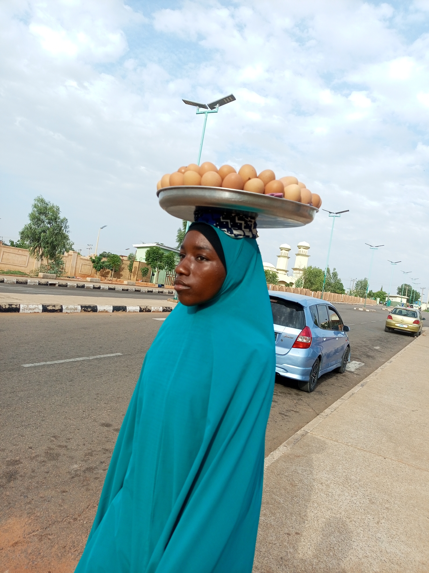 In the city of Gombe, Nigeria, a young woman wearing a turquoise hijab and traditional clothing walks along a paved sidewalk next to a road. She is balancing a large metal tray of eggs on her head. The tray is filled with numerous brown eggs. Behind her, a light blue hatchback car is parked on the road, and further down the road, a gold sedan is visible. The environment is a typical urban scene with a road, sidewalk, and buildings in the background. The sky is overcast with scattered clouds, and there are streetlights visible along the road.