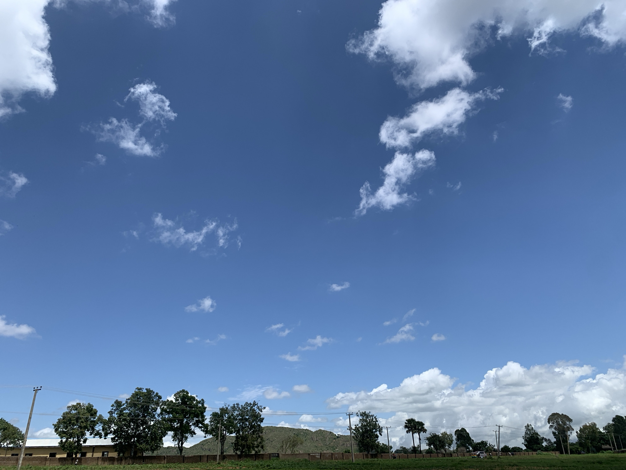 A wide, bright blue sky fills the upper portion of the image, dotted with scattered white cumulus clouds. Below the sky, a low horizon line is visible, featuring a dense line of trees with dark green foliage. A light brown building with a dark roof is partially visible behind the trees on the left side of the frame. Power lines stretch horizontally across the scene, supported by telephone poles. A brick wall runs along the ground in front of the building and trees. In the background, a large, verdant hill rises behind the trees, with a few scattered clouds hovering near its peak. The overall environment appears to be a rural or semi-rural landscape.