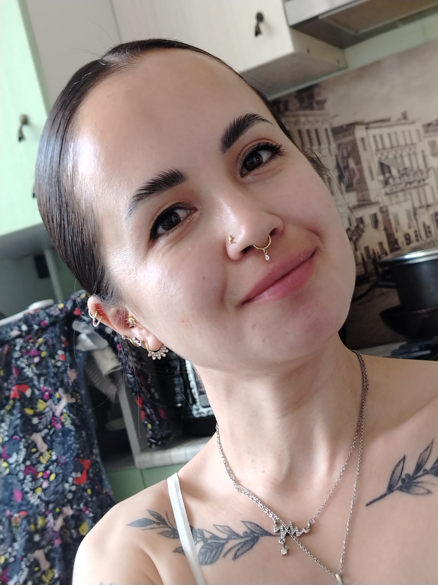 A close-up photograph shows a woman with dark hair pulled back, exposing a smooth forehead. She has dark eyes and well-defined eyebrows. The woman has a gold nose ring and multiple earrings in her left ear. She wears a silver necklace. Tattoos are visible on her chest and shoulder. In the background, a kitchen scene with a patterned wall covering, cabinets, and a cooking pot are visible. The setting appears to be indoors, in a domestic environment. No information about the location in the city of Noviy, Russia can be inferred.
