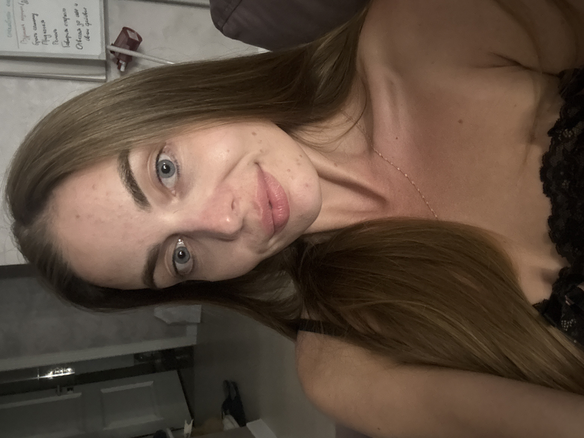 The image shows a young woman with long, light brown hair and blue eyes. She is lying down, with her head resting on her arm. Her gaze is directed upwards, and she has a slight smile on her face. She appears to be indoors, in what looks like a bedroom or living area, given the soft lighting and furnishings in the background. The time of day is not clearly discernible, but the lighting suggests it could be evening or nighttime.
There is text visible on a whiteboard in the background, which appears to be written in Cyrillic script. While the specific words are not fully legible, some phrases that can be partially made out include "Прически", "Свадебные", "Народные", "Подарки", and "Отвечая за свой". These words relate to hairstyles, weddings, gifts, and responsibilities, suggesting the whiteboard may contain notes or a to-do list related to event planning or personal services.
The woman is wearing a black top with lace detailing. A delicate gold necklace is visible around her neck. The overall mood of the image is relaxed and intimate. The location context of Sochi, Russia, is provided, though there are no specific visual cues in the image to confirm this location.