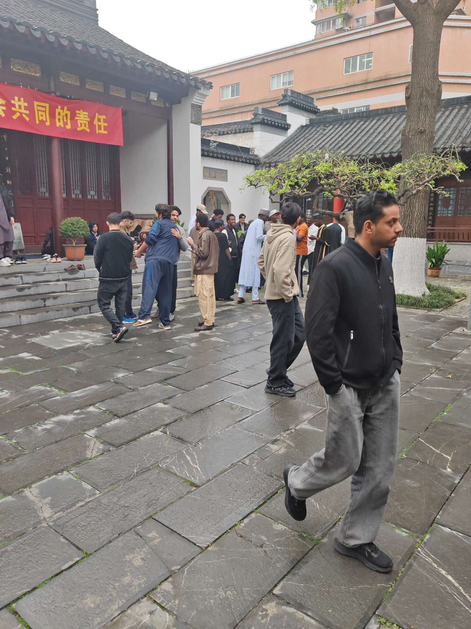 A medium-wide shot shows a group of people in the courtyard of the Nanjing Mosque in the Qinhuai District of Nanjing, China. The courtyard is paved with large stone tiles that appear wet. A man in a black jacket and gray pants walks toward the right of the frame, his hands in his pockets. Several other men stand in small groups near the steps leading up to the main building, a traditional Chinese-style structure with a red banner hanging above the entrance. Some of the men are dressed in traditional Islamic clothing. A few shoes are left on the steps of the building. In the background, there's a two-story building with light-colored walls and a traditional Chinese roof. A tree is located in the courtyard and in the background.