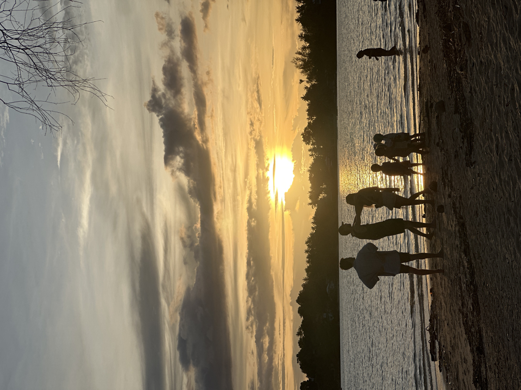 The image depicts a serene sunset scene on a beach, likely in ตำบลเกาะพะงัน, Thailand, given the lush tropical foliage lining the distant shore and the warm, golden light. The sky is a canvas of dramatic clouds, with the sun breaking through, casting a radiant glow across the water and illuminating the scene.
In the foreground, a sandy beach is sparsely littered with natural debris such as dried leaves and driftwood. Several individuals are silhouetted against the bright sky and water. On the left, three adults stand near the water's edge, their figures dark against the backdrop. One appears to be facing away, hands on hips, while the other two are in conversation.
Further to the right, closer to the water, a group of children, also silhouetted, are engaged in playful activity, some wading in the shallow water. A lone figure stands further out in the water on the right side of the frame, their posture suggesting they are observing the scene or enjoying the water.
The water itself is calm, with gentle ripples reflecting the warm hues of the sunset. The line of trees on the horizon creates a dark, solid silhouette, emphasizing the vibrant colors of the sky. The overall atmosphere is one of tranquility and natural beauty, with human presence adding a touch of life to the picturesque landscape.