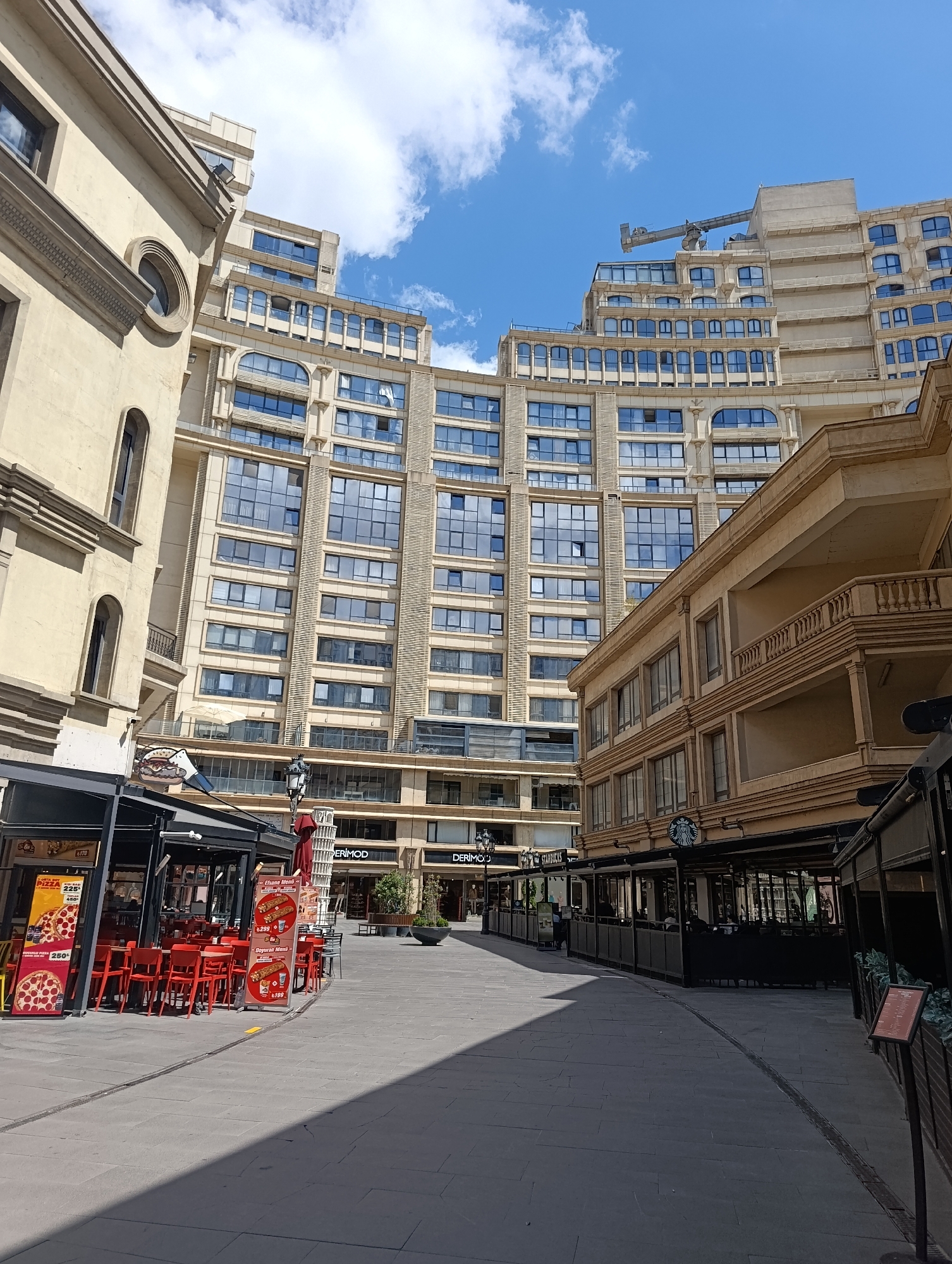 The image depicts an outdoor pedestrian walkway lined with commercial establishments and modern multi-story buildings in Gaziosmanpaşa, Turkey. The walkway is paved with large gray tiles, with a drainage channel running along its center.
On the left side of the walkway, a restaurant with a black awning displays red tables and chairs. Signage for "Pizza" is visible, along with menu boards featuring pizza and other food items. To the right of the restaurant, a tall, slender structure resembling the Leaning Tower of Pisa, but seemingly a decorative element or advertisement, stands vertically.
Further along the walkway to the right, another commercial area features a Starbucks with an outdoor seating area enclosed by dark screens. The iconic Starbucks logo is prominently displayed. Behind these commercial fronts, several large, modern buildings rise. These buildings feature a light beige facade, numerous windows with blue-tinted glass, and intricate architectural details such as arched window frames and decorative vertical elements. The buildings appear to be a mix of residential and commercial spaces, with a construction crane visible atop the tallest structure in the background.
The sky above is blue with scattered white clouds. The scene appears to be a public space, likely a commercial district or a modern development area within the city. No individuals are clearly identifiable or interacting in the scene, suggesting a moment of relative quiet or distance. The lighting suggests it is daytime, with shadows cast by the buildings and awnings onto the walkway.