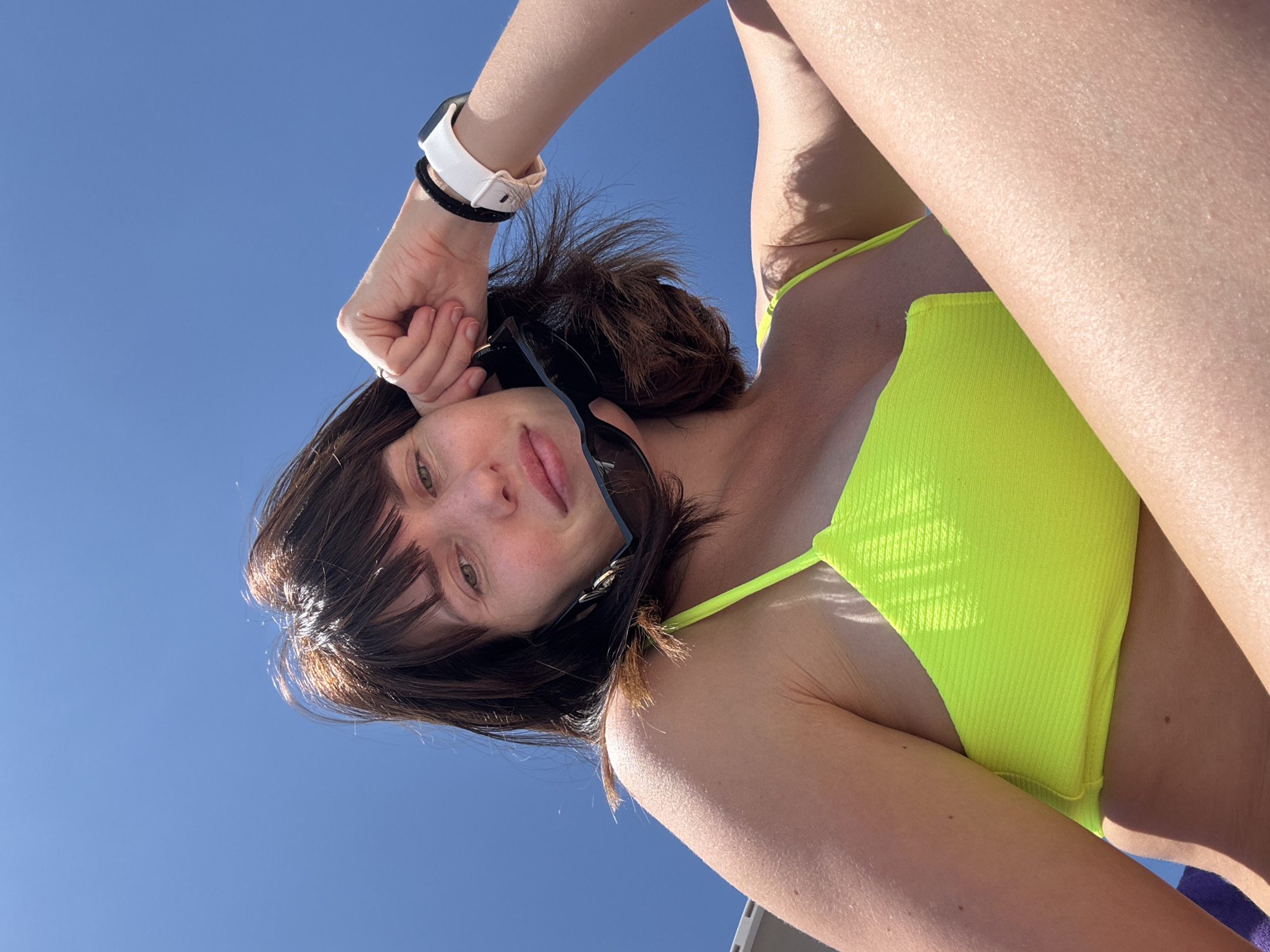 The image shows a close-up, low-angle selfie of a young woman against a clear blue sky. She has dark brown hair, fair skin, and is wearing a bright neon yellow bikini top. Her left arm is raised, with her hand resting near her forehead, obscuring part of her face. She is wearing sunglasses pushed up onto her hair and a white and black fitness tracker on her left wrist. The lighting suggests it is daytime, likely sunny. The background is dominated by the bright blue sky. The image appears to be taken outdoors, possibly at a beach or a location with an open sky. The overall mood is casual and summery.