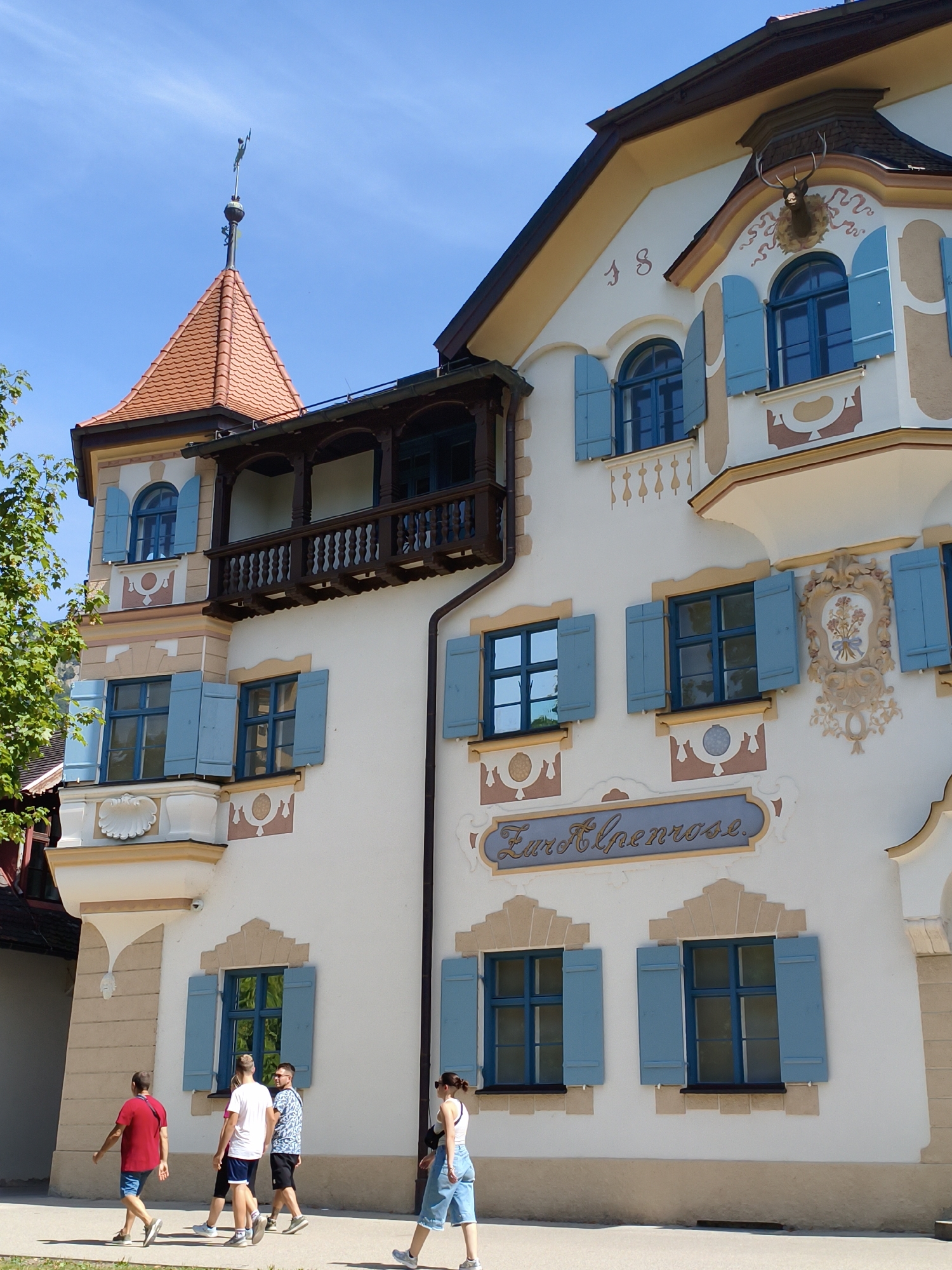 The image shows the facade of a traditional Bavarian building in Schwangau, Germany, identified by the sign "Zur Alpenrose" above two arched windows. The building is painted white with light brown and tan decorative accents. It features several blue shuttered windows, some of which are arched. A wooden balcony with decorative railings juts out from the upper story. A red-tiled conical roof with a weathervane sits atop a smaller tower-like section of the building. A deer head trophy is mounted above a window on the upper right of the facade. In front of the building, on a paved walkway, four people are walking: a man in a red shirt and blue shorts, a man in a white t-shirt and blue shorts, a man in a patterned shirt and black shorts, and a woman in a white top and denim shorts. The sky is clear and blue.