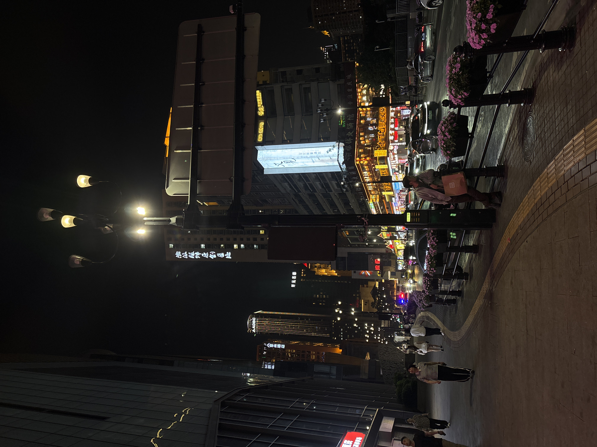 This is a night shot of a street scene in Yuzhong District, China. The scene is oriented vertically. Buildings flank both sides of the street, illuminated by lights and neon signs. A few high-rise buildings are visible in the distance. On the left side of the image, there's a building with the visible text "偷派服饰原創基地", "AADBC", "亚朵酒店", "开元名都", "又见观酒店", and "命格饼火锅馆专注重庆味". People can be seen walking on the sidewalk. Planters with purple flowers line the street. Cars are visible in the distance, with their headlights on. Street lamps provide additional illumination.
