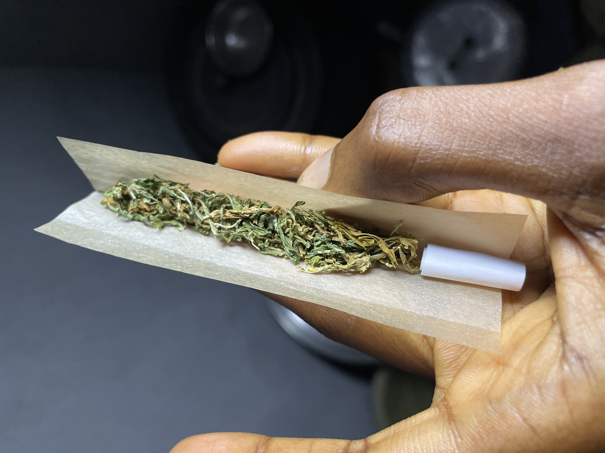 A close-up shot shows a person's hand holding a marijuana joint and rolling paper. The joint is partially rolled and contains dried cannabis. A white filter tip is visible at the bottom of the joint. The background is dark and blurred, suggesting an indoor setting. The image appears to be taken in Nkpor, Nigeria.