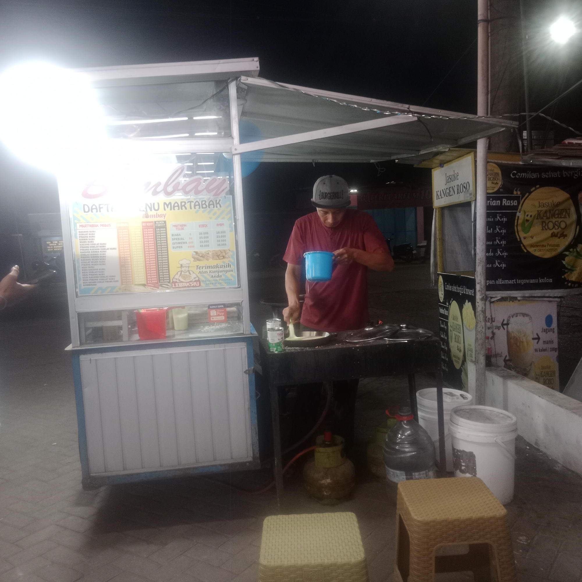 The image depicts an outdoor scene at night in Tegowanu Kulon, Indonesia, likely a street food stall. A man wearing a maroon t-shirt and a grey baseball cap is standing behind a food cart and a cooking station, holding a blue cup and appearing to be in the process of cooking. The food cart has a menu board displaying "DAFTAR MENU MARTABAK" and various prices for different martabak flavors. Next to the cart, there is a cooking stove with a pan, a gas cylinder, and several large plastic containers. In the background, there are signs with text in Indonesian, including one that says "Jasuke KANGEN ROSO" and another advertising "Cemilan sehat bergizi". The ground is paved with brick-like tiles. The overall lighting suggests artificial light sources are being used.