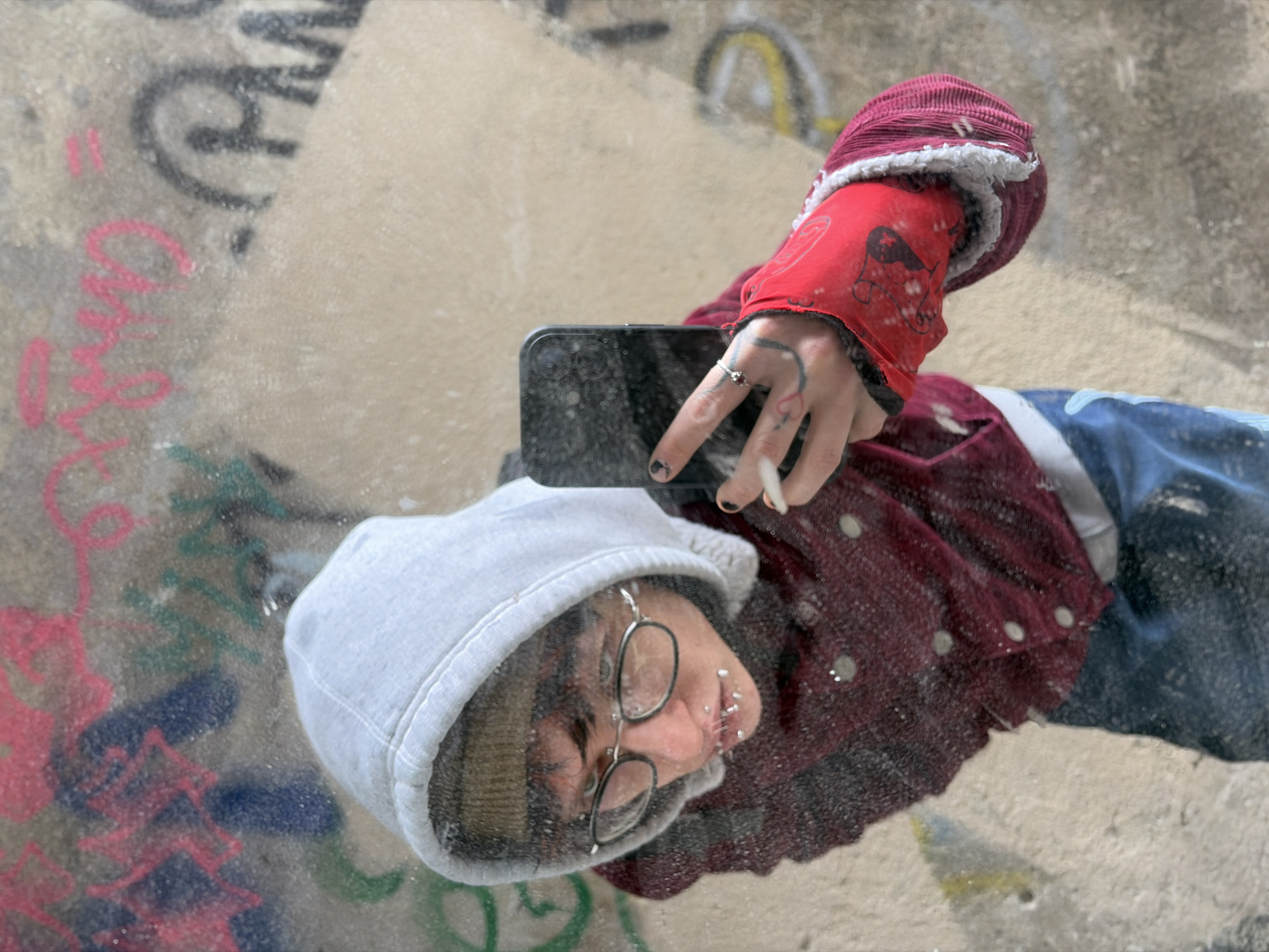 A person is reflected in a glass surface, possibly a window or a mirror, with graffiti visible in the background and on the glass itself. The person is wearing a light gray hooded sweatshirt, glasses, a maroon corduroy shirt or jacket, and blue jeans. Their left hand, which has tattoos, is holding a black smartphone, and they are holding a lit cigarette between their index and middle fingers. The background features various colors of graffiti on a wall. The setting may be in Sofia, Bulgaria, but without further information this cannot be verified. The reflection gives the impression that the person is upside down. The glass surface appears to be smudged and marked, obscuring some of the details.