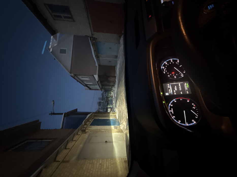 A view from inside a car, at dusk or dawn, looking down a narrow alley. Buildings line both sides, with garage-like doors visible on the right. The sky is a deep blue. The car's dashboard and instrument panel are visible, with illuminated gauges. The vehicle appears to be stationary or moving very slowly.