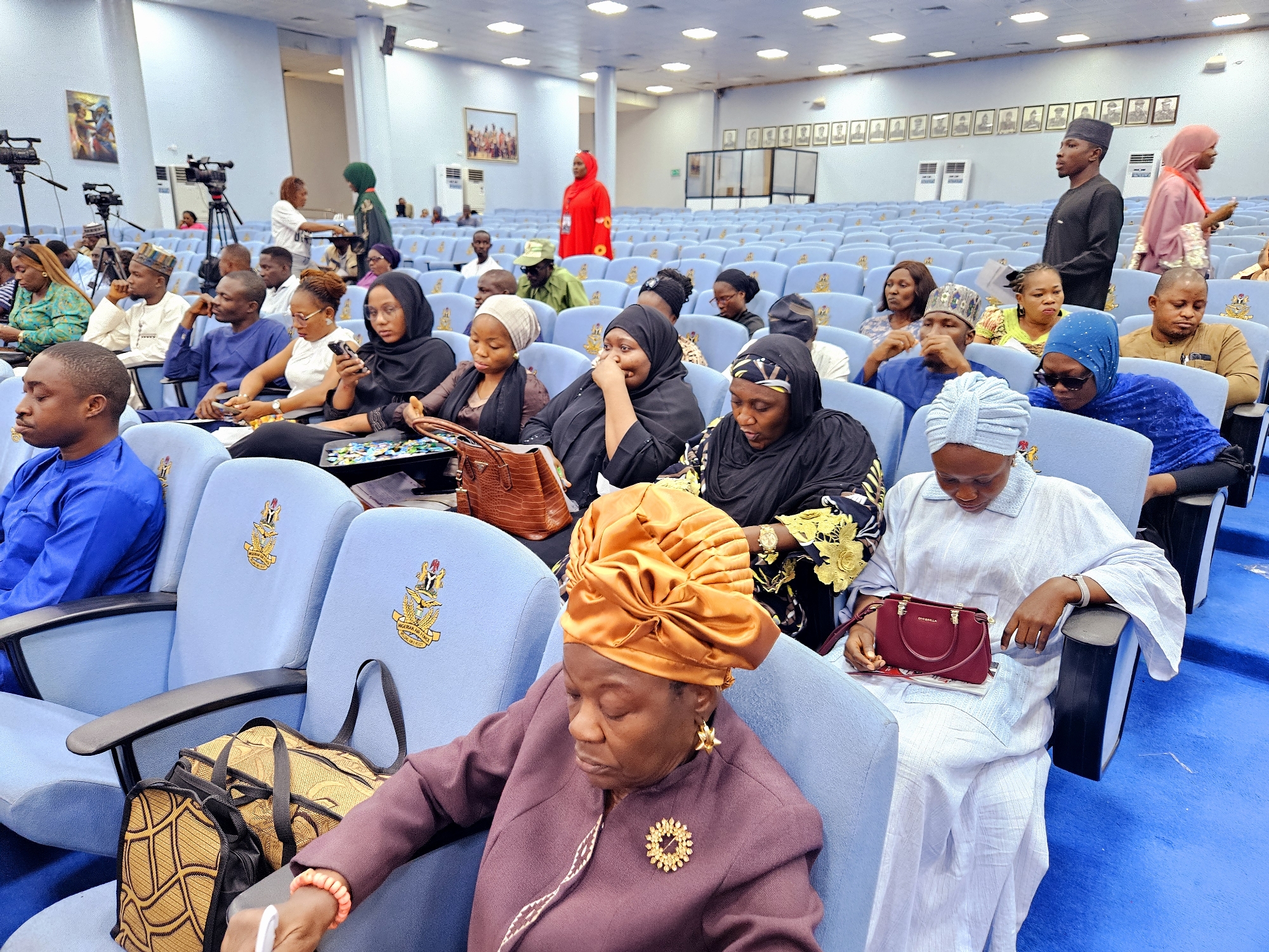 The image shows an indoor gathering in a hall with rows of light blue seats. The seats have an emblem on them. Several people are seated in the foreground and middle ground. In the foreground, an older woman wearing a maroon jacket and an orange headwrap is seated, looking down. To her right, a woman in a white outfit is seated, holding a small red purse and looking at her phone. Behind them, more people are seated, many wearing headscarves. In the background, there are rows of empty seats and a stage area with cameras and microphones. A person in a red outfit is standing near the stage. The overall environment appears to be a conference room or auditorium. The presence of cameras and a formal seating arrangement suggests an event, possibly a political or official gathering. The location is Gwarinpa, Nigeria, as indicated in the prompt.