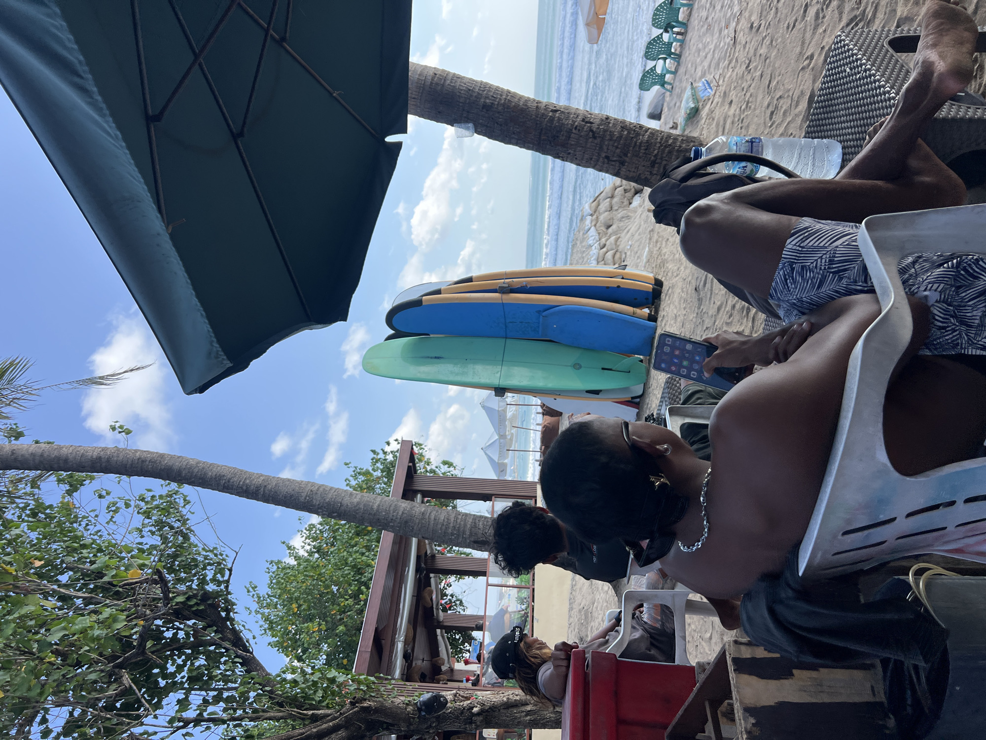 The image captures a beach scene in Kuta, Indonesia, likely a popular surfing spot given the presence of surfboards. In the foreground, a person is seated in a white plastic chair, looking at their phone. They are wearing patterned shorts and a necklace. To the left, another person is also seated, wearing a black baseball cap. Behind them, more people are visible, as well as a wooden structure that appears to be a beach bar or restaurant. Several surfboards of varying colors stand upright near the water's edge. The ocean stretches to the horizon under a partly cloudy sky. The beach is sandy, with some beach chairs scattered in the background. A large green umbrella provides shade. The overall atmosphere suggests a relaxed day at the beach, with people enjoying the sun, sand, and surf.