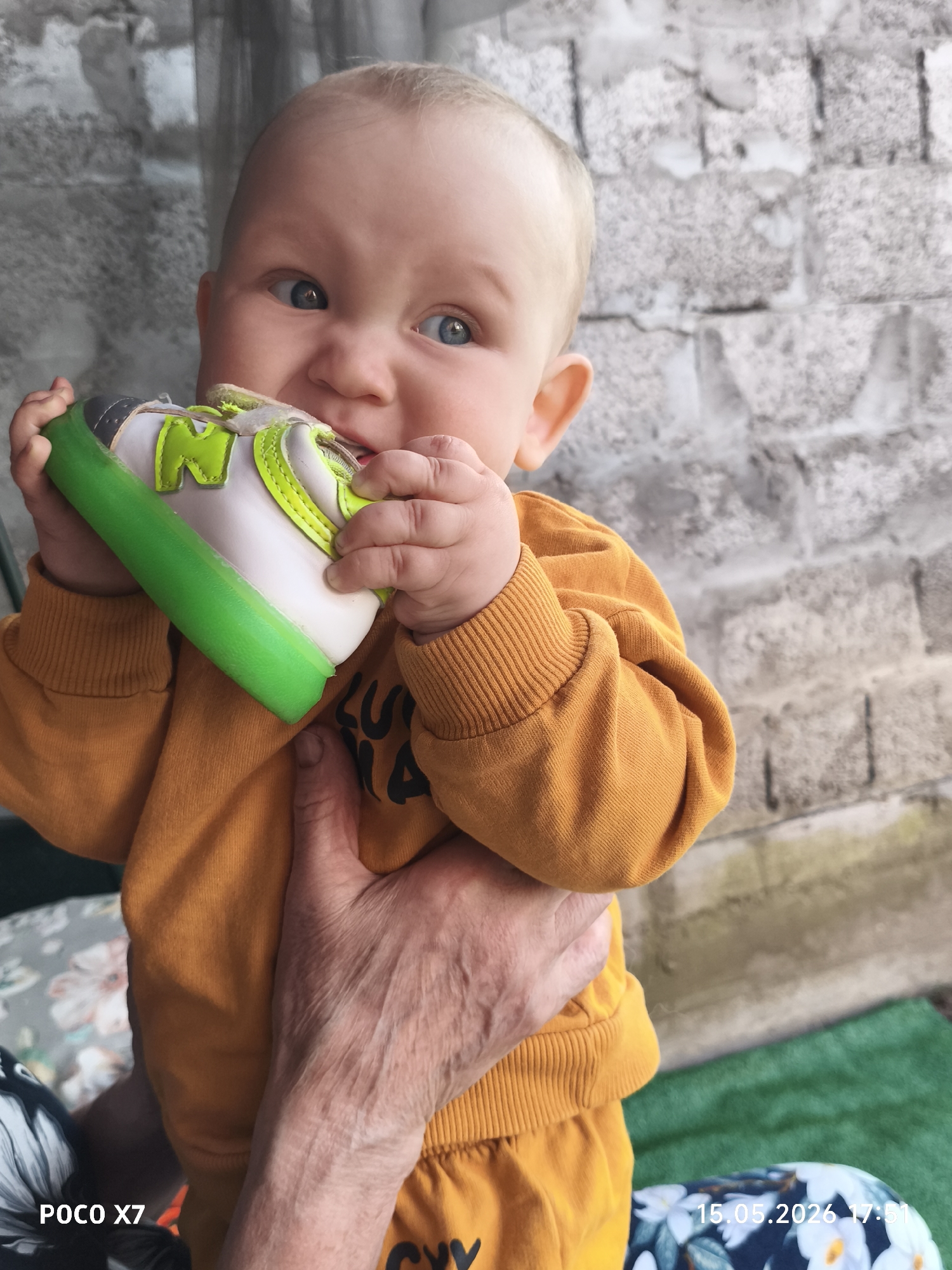 A baby with fair skin and blue eyes is being held in an adult's hand. The baby is wearing an orange sweatshirt and matching pants. The baby is chewing on a shoe that is white with green trim and a large letter "N" on the side. The background is a gray brick wall. The location is Chaikovsky, Russia. The photograph was taken on May 15, 2026, at 17:51, using a POCO X7 camera. The baby's expression is intense as they chew on the shoe. The hand holding the baby appears to be older, based on the visible wrinkles. There is a floral patterned fabric visible in the lower left corner, possibly part of a cushion or clothing. There is a green surface below the floral fabric.