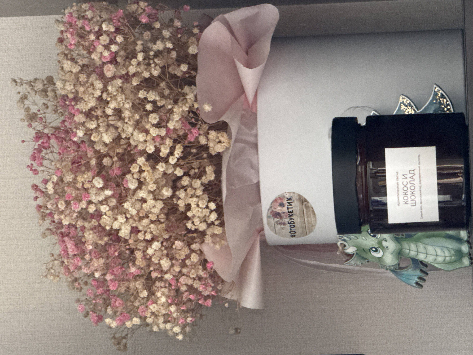 A still life composition features a bouquet of dried gypsophila flowers, predominantly in shades of pink and white, arranged in a white cylindrical box. A pale pink tissue paper lining is visible at the top of the box. A round sticker with a wooden texture and floral motifs is affixed to the side of the box, bearing the hashtag "#огобукетик". In front of the box, to the left, a small, green, cartoon-like dragon figurine with blue wings is positioned. To the right of the dragon and slightly behind it, a dark glass jar with a black lid is visible. A white label on the jar displays Cyrillic text that reads "КОКОС И ШОКОЛАД" (Coconut and Chocolate), indicating it contains an aromatic candle. Behind the jar, a stylized, light-colored Christmas tree ornament with sparkling embellishments is partially visible. The background appears to be a neutral, textured surface, possibly a wall. The overall scene is presented indoors, with soft lighting. There is no indication of this image being taken in Nizhny Novgorod, Russia, or any specific city.
