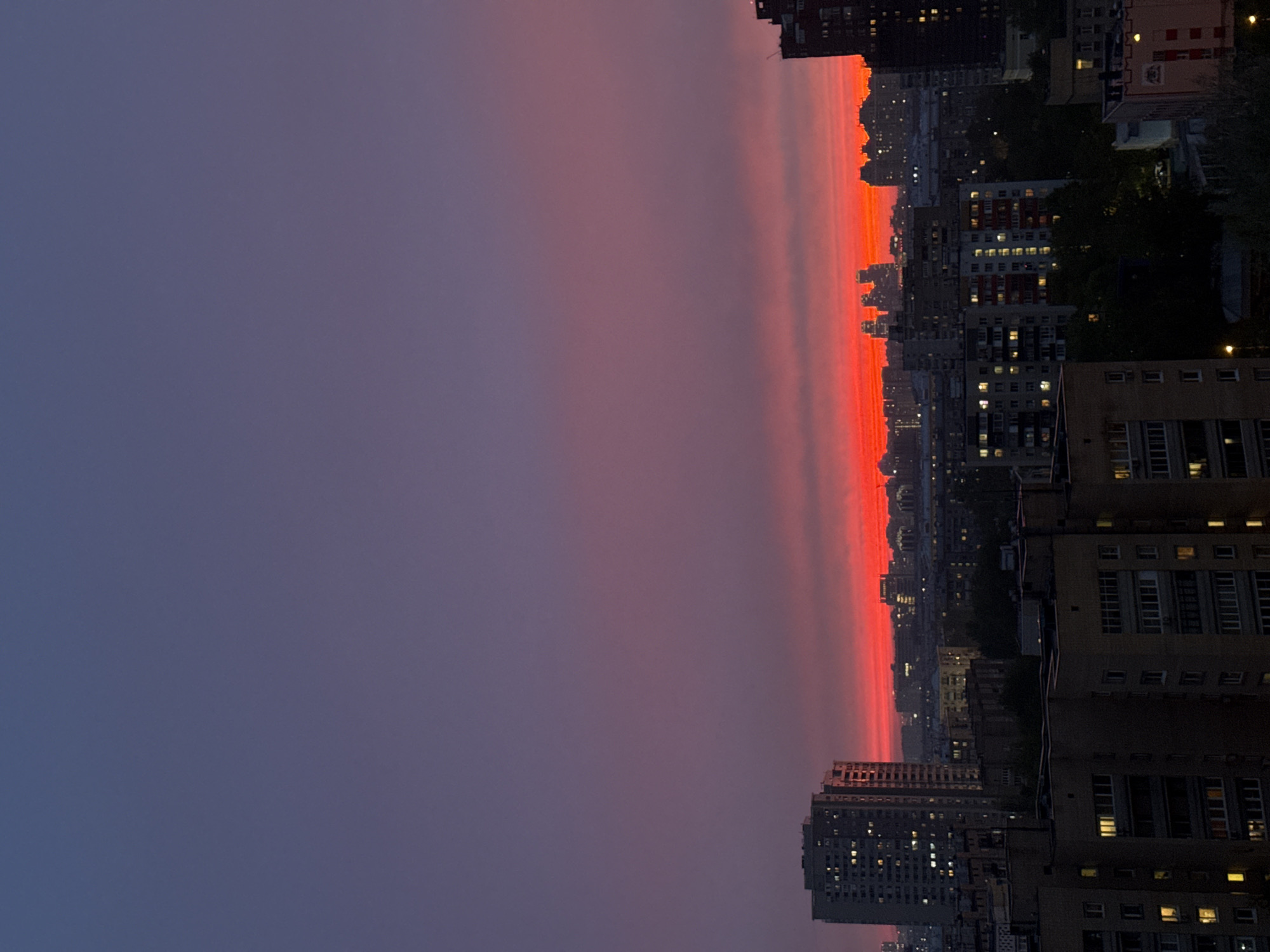 The image shows a cityscape at sunset or sunrise, likely in Moscow, Russia. The sky transitions from a dark blue-purple on the left to a vibrant red-orange along the horizon, creating a dramatic light effect. Multiple tall buildings and apartment complexes are silhouetted against the bright sky. Some windows are lit, indicating activity inside. Trees are visible among the buildings, adding a touch of greenery to the urban landscape. The image is rotated counterclockwise.