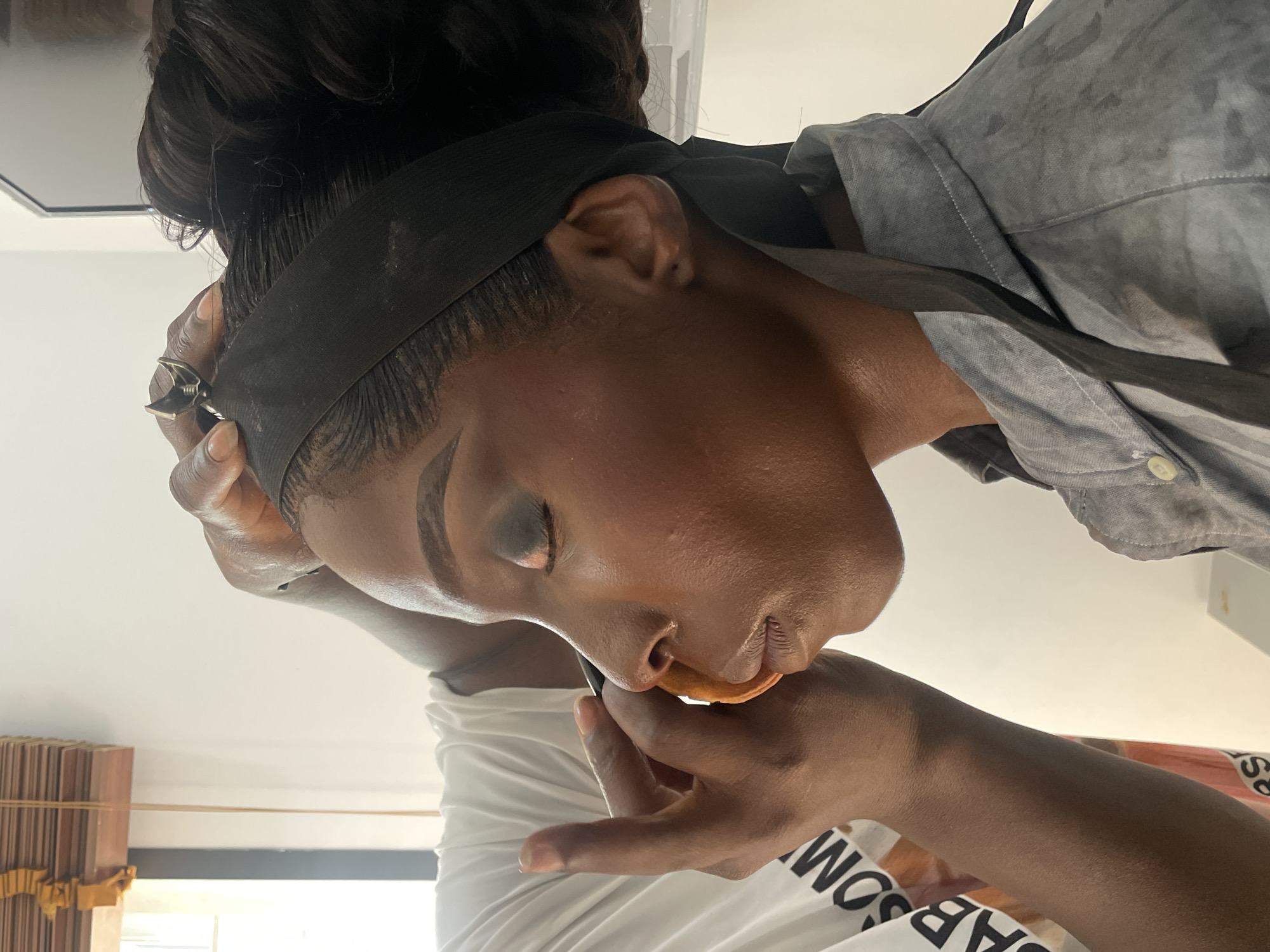 A close-up shot, likely taken indoors, shows a dark-skinned person with a black band around their forehead and a makeup artist applying product to their face. The person receiving makeup has their eyes closed and appears to be leaning their head back slightly. The makeup artist is holding a makeup sponge or brush. A gray collared shirt is visible on the right side. The location of Jos, Nigeria is not clearly evident from the image alone.
