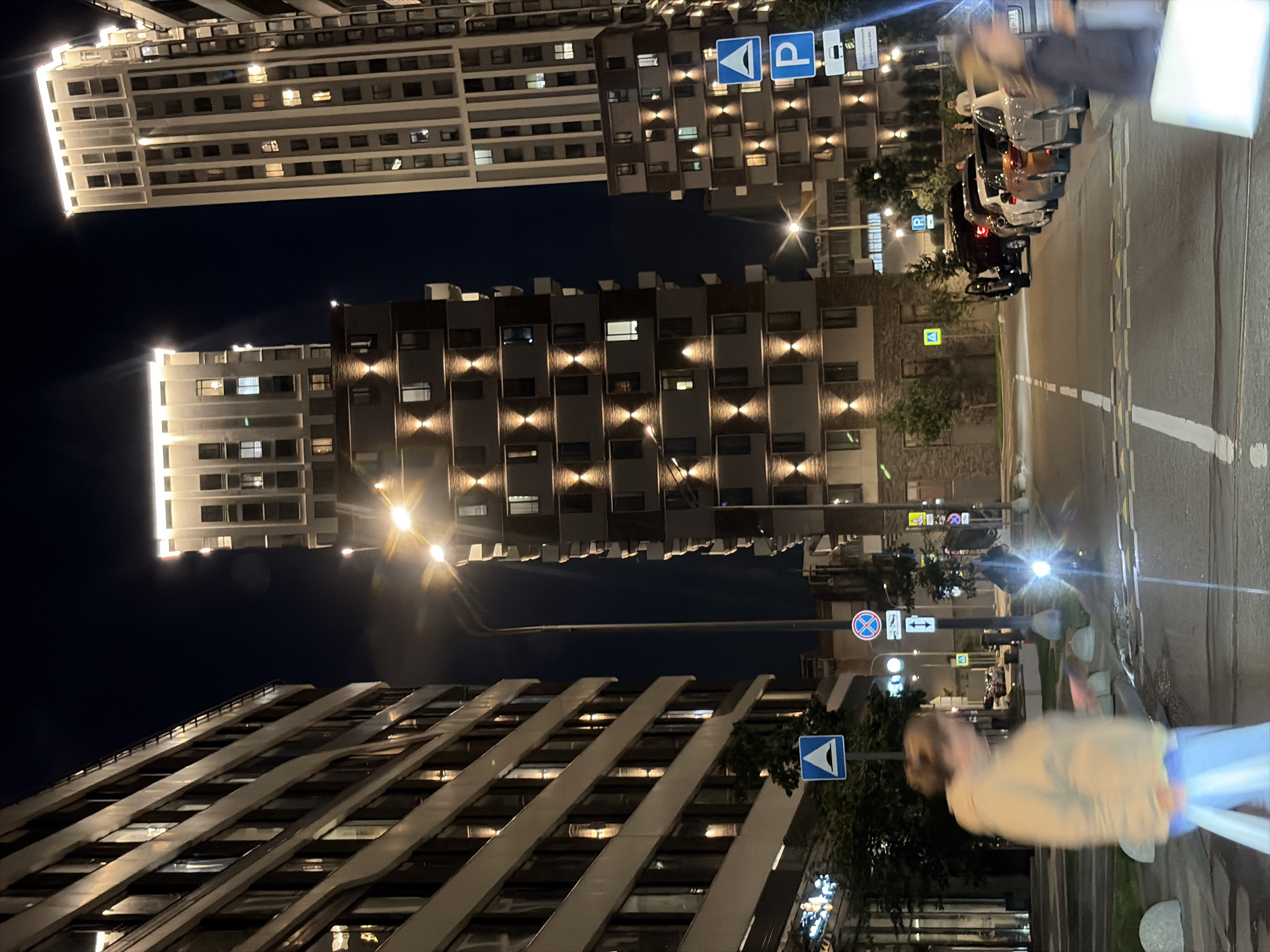 This is a nighttime exterior shot of a modern residential complex in Moscow, Russia. The image captures a street scene with several tall, contemporary apartment buildings illuminated by architectural lighting. The buildings feature a grid of windows and balconies, with accent lighting highlighting their facades.
In the foreground, a woman in a tan hoodie and blue pants is walking past the camera, appearing slightly blurred due to motion. Further down the street, another person is visible on what appears to be a scooter or motorcycle, with its headlight illuminated. Several cars are parked along the right side of the street, and various traffic signs are visible, including a blue parking sign, a pedestrian crossing sign, and a circular red and blue sign indicating a restriction.
The road is paved and marked with white lines, and there are some speed bumps visible. Streetlights and the lights from the buildings create a well-lit, urban nighttime atmosphere. The sky is dark, indicating it is night.