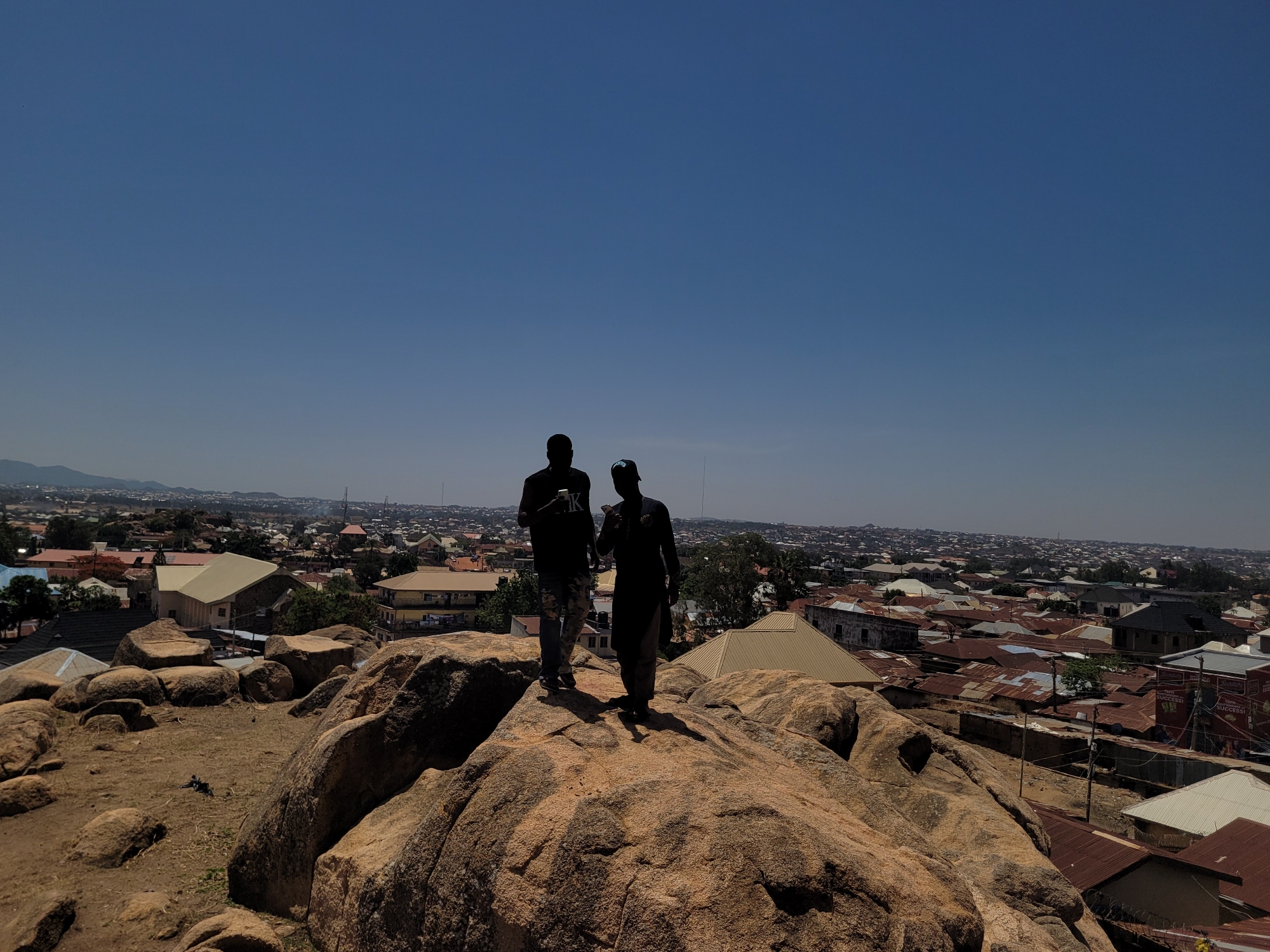 Two people stand on top of a large rock formation, overlooking a dense urban landscape under a clear blue sky. The individuals are dark silhouettes. One is wearing a t-shirt with a large letter "K" on it, holding a phone. The other is wearing a traditional muslim cap, also holding a phone. The city of Jos, Nigeria, stretches out in the background, showing densely packed buildings with primarily corrugated metal roofs. The foreground is characterized by the rough, exposed surfaces of the large rocks.