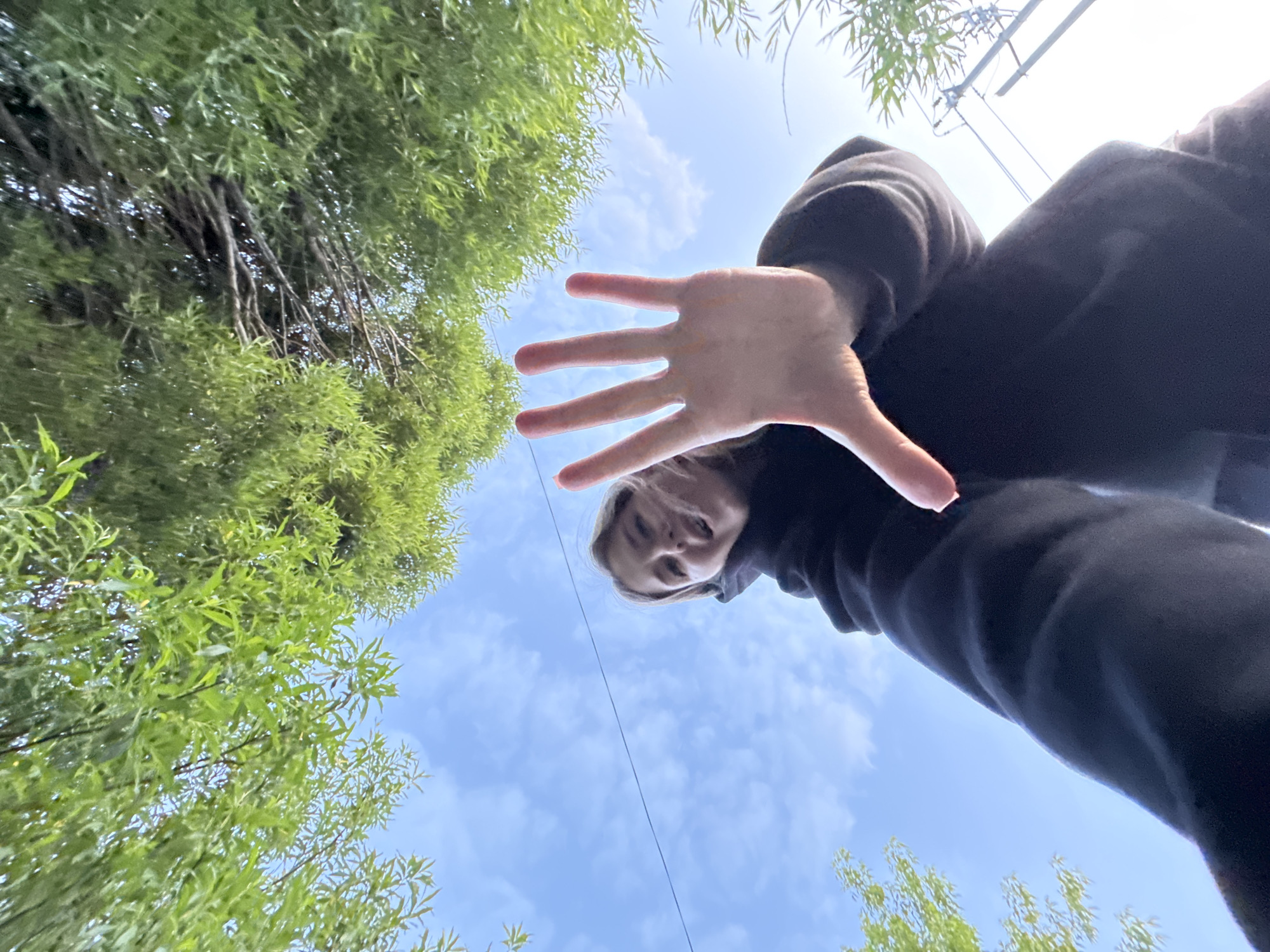 A low-angle shot looking up at a person wearing a dark hooded sweatshirt with their hand outstretched towards the camera. The person's face is partially visible. The background consists of trees with green leaves and a blue sky with some clouds. A power line is also visible in the sky. It is not possible to confirm the location is in the city of Горный, Russia from the image.
