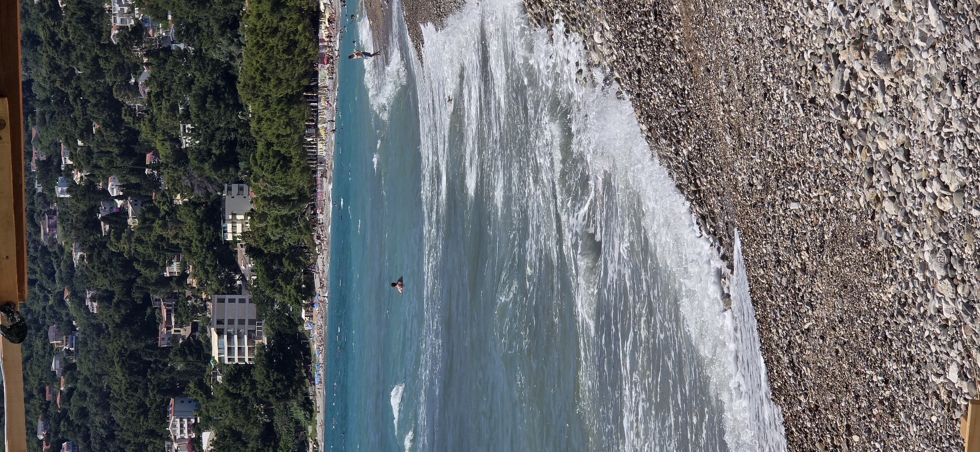 The image depicts a pebble beach in Bar, Montenegro, with the Adriatic Sea in the foreground and a densely forested hillside dotted with buildings in the background. The water is a vibrant turquoise and exhibits small waves crashing onto the shore. Several individuals are visible in the sea, with one person swimming and another standing near the water's edge. The beach itself is covered in a variety of grey and brown pebbles. In the distance, buildings are nestled among lush green trees on a hillside. The sky is clear and blue. A wooden structure is partially visible at the top of the frame.