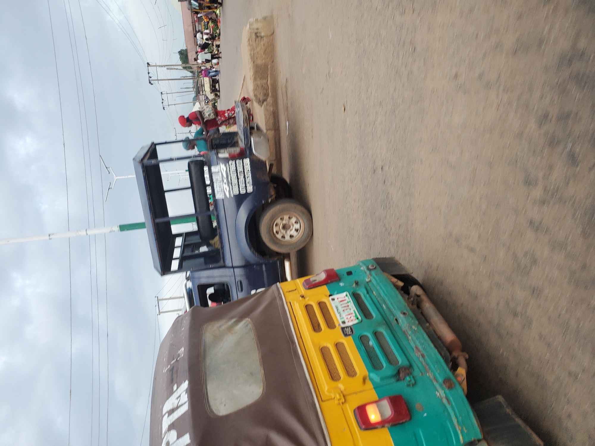 The image depicts a street scene in Bukuru, Nigeria, under an overcast sky. In the foreground, a yellow and green auto-rickshaw, also known as a Keke Napep, is angled towards the left. Its license plate, visible on the rear, reads "KSR 314 LZ". To the right of the Keke Napep, a dark blue pickup truck with a covered bed is parked. Two individuals, a woman wearing a red headscarf and a teal-colored top, and another woman with a red head covering and a patterned skirt, are standing near the pickup truck, appearing to be engaged in conversation or observing the surroundings. In the background, a market is visible with stalls and people moving about. Several utility poles and electrical wires are present, indicating an urban environment. The road surface is paved and appears to be dusty.