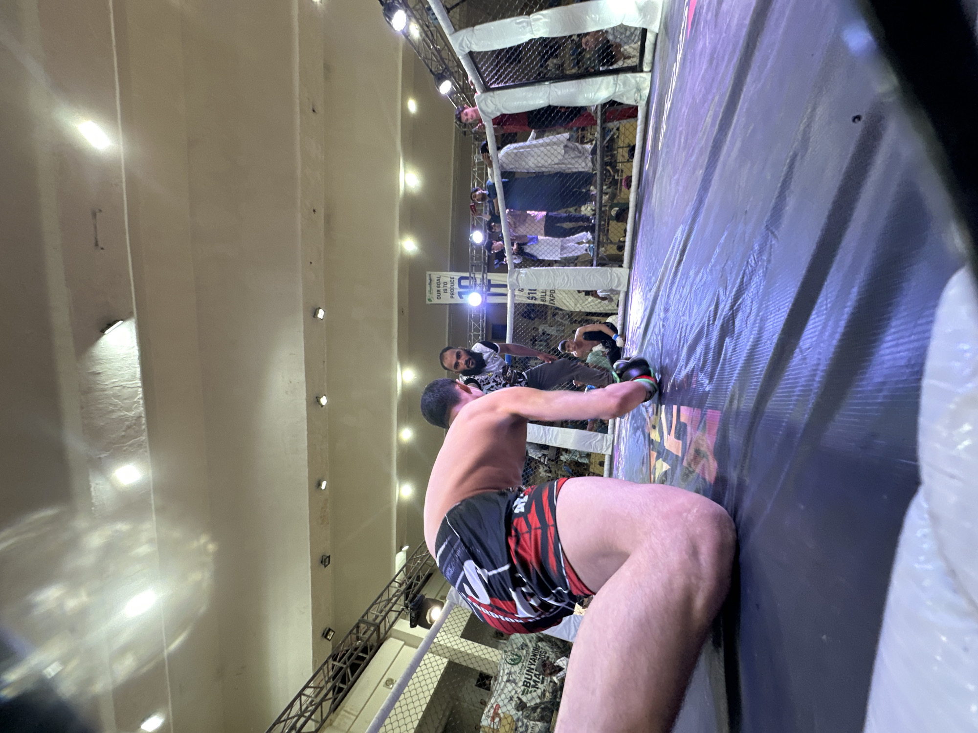 A mixed martial arts fight is taking place in a cage in Quetta City Tehsil, Pakistan. A fighter is pressed against the cage wall. He is wearing black and red shorts and gloves. Another man stands near the cage watching. People are in the background looking at the event. The arena has a high ceiling with rows of lights.