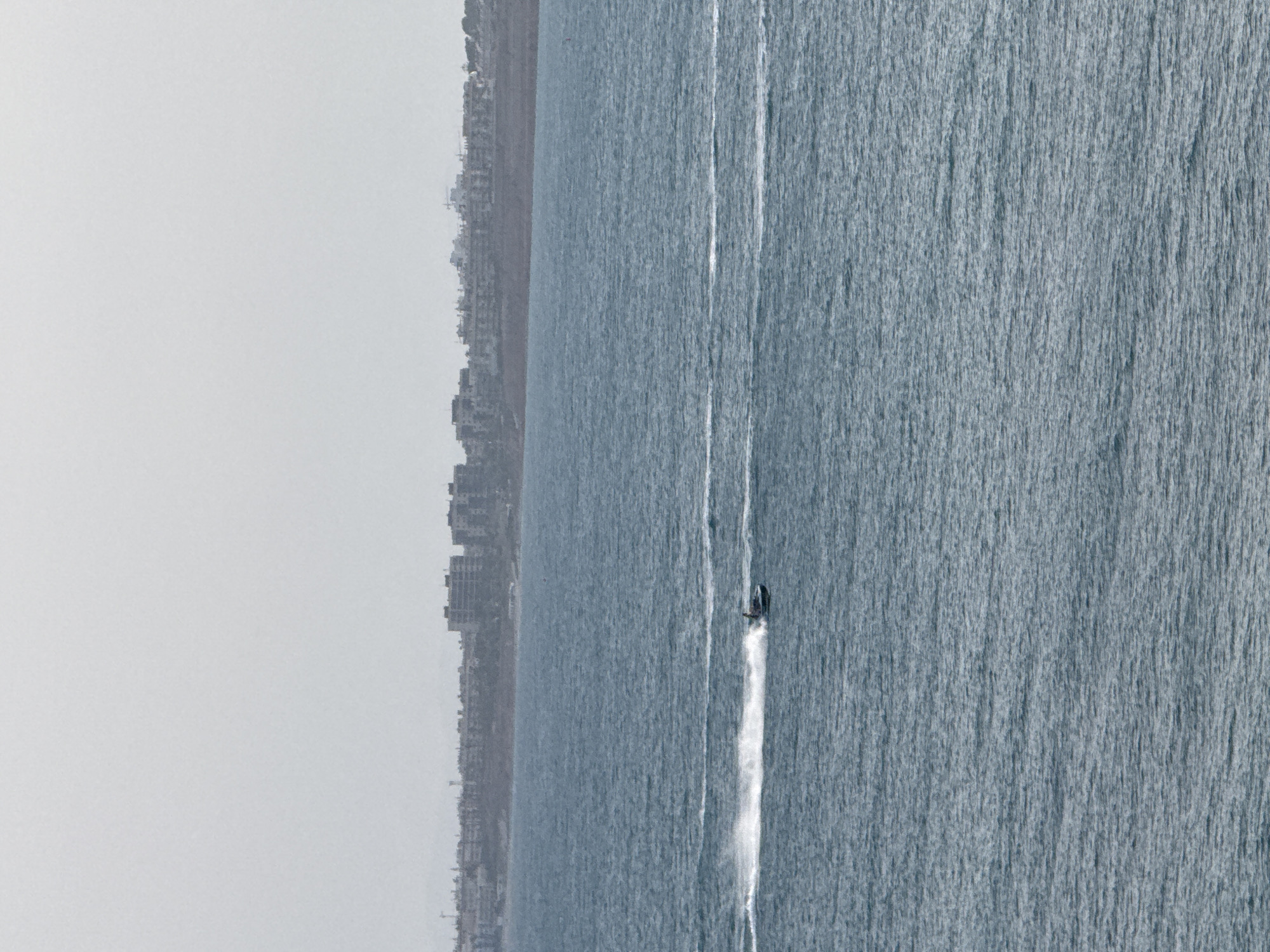 A jet ski is being operated on a choppy, blue ocean. The jet ski is leaving a white spray in its wake. In the background, a distant city skyline is visible under a hazy, overcast sky. The city appears to be located on a coastal plain.