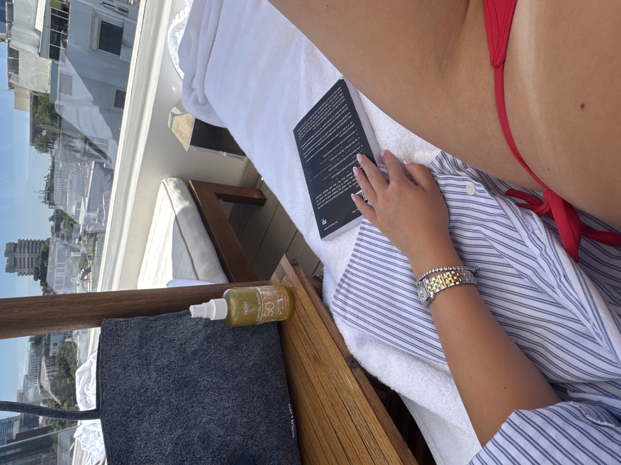 This is a medium shot, outdoor, eye-level photograph taken on a sunny day in Tel Aviv, Israel. The scene depicts a person lounging on a white towel on a wooden deck chair. The person is wearing a red bikini bottom and a blue and white striped shirt. They are wearing a watch with a silver band and a diamond bracelet. Their left hand, with manicured nails, is resting on the towel. A black book with text in Hebrew is open on the towel next to the person's hand. A grey canvas tote bag, a bottle of Vichy Capital Soleil SPF 50 sunscreen, and a white folded towel are on the wooden deck chair. The background shows a cityscape with buildings and trees, as seen through a reflection on a glass barrier.