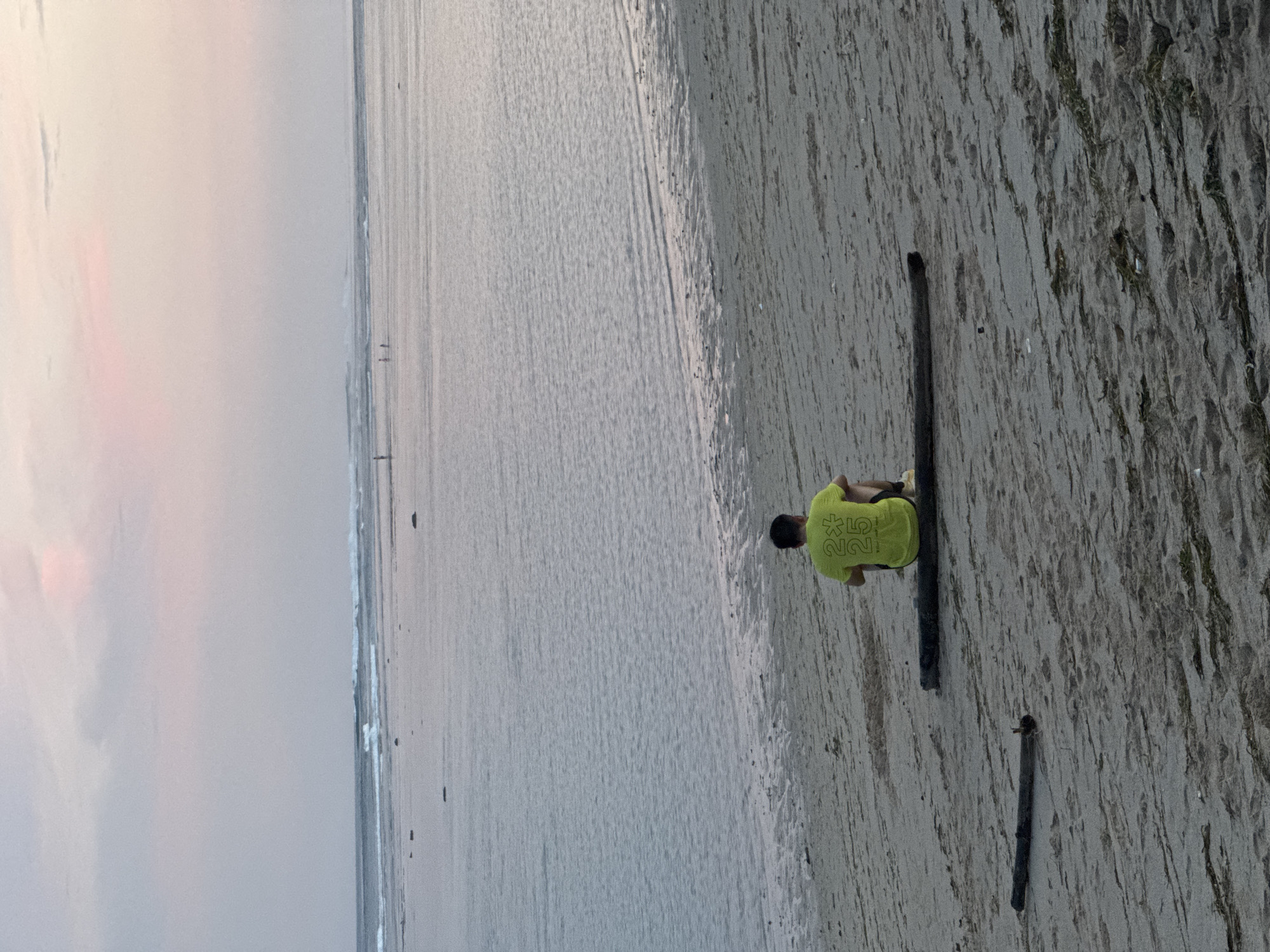 The image depicts a tranquil beach scene at either dawn or dusk, set in Kuta Selatan, Indonesia. The photograph is oriented vertically, showcasing a wide expanse of sandy beach meeting the ocean under a soft, diffused sky with subtle hints of pink.
In the foreground, a lone individual is seated on a dark, weathered log, viewed from behind. The person is wearing a bright, neon yellow-green t-shirt. Visible on the back of the shirt are the digits "*25*", accompanied by a star or asterisk-like symbol. Another smaller piece of wood or log lies on the sand nearby.
The vast sandy beach stretches towards the calm ocean, where gentle waves are visible near the shore. The expansive horizon is somewhat obscured by a hazy atmosphere. In the far distance, faint figures of what appear to be other people are barely discernible along the waterline. The scene conveys a sense of peaceful solitude and contemplation, with the person seemingly observing the sea or resting.